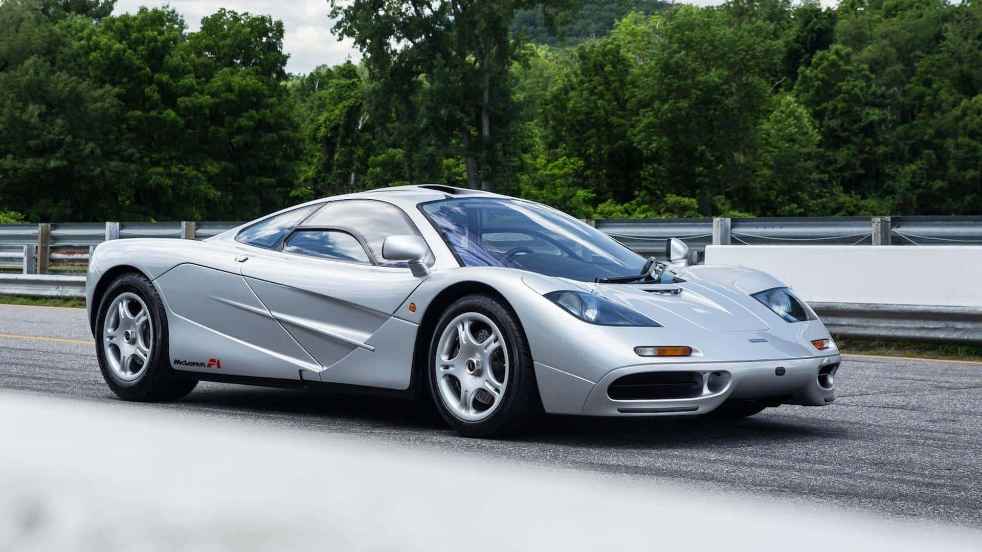 Mclaren F1 桌布 壁紙 — 一架流線型銀色McLaren F1超級跑車沿著蜿蜒的道路行駛，兩旁是茂密的綠樹和金屬護欄。