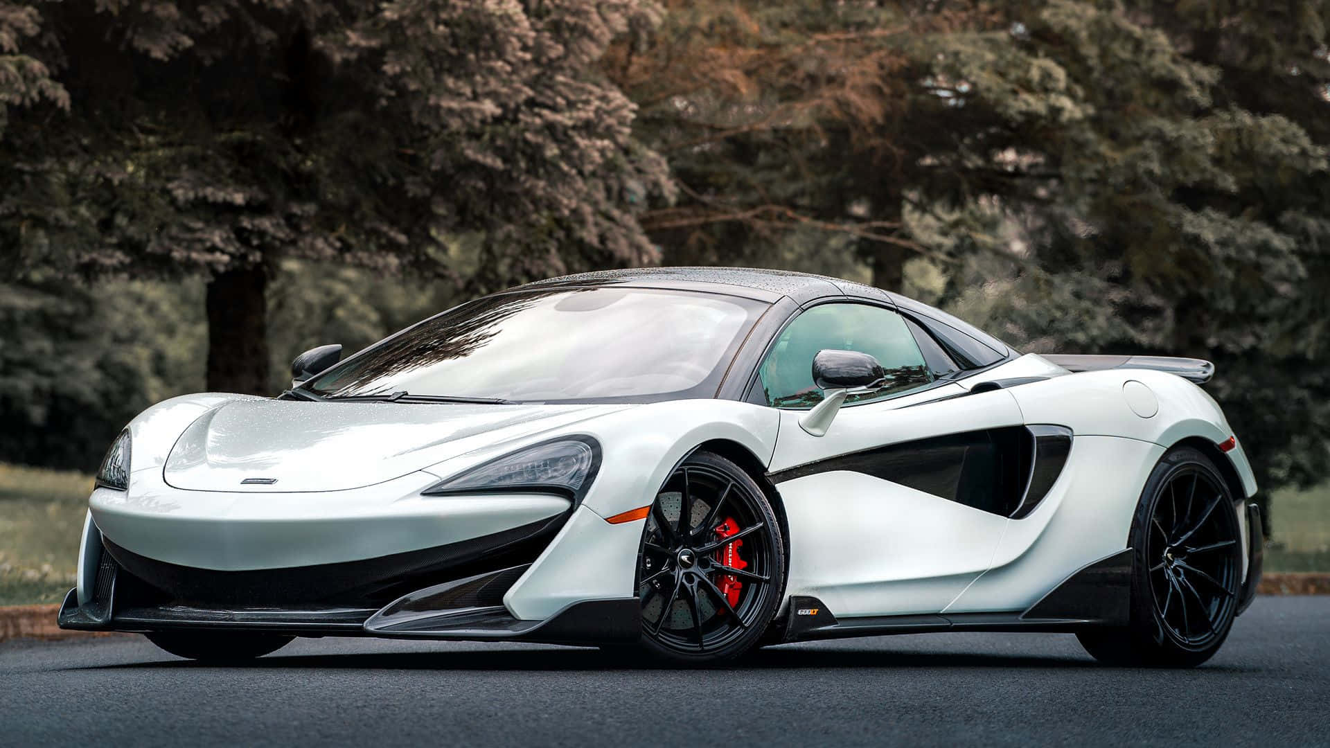 Mclaren 600lt 桌布 壁紙 — 一輛時尚的銀色McLaren跑車，帶有黑色飾件和紅色制動卡鉗，停在樹木環繞的瀝青路面上。