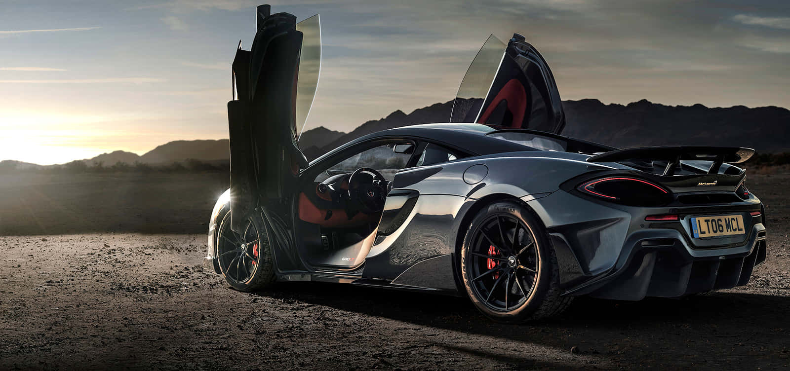 Mclaren 600lt 桌布 壁紙 — 一架線條流暢的銀色McLaren跑車，鷗翼車門打開，在日落時分的沙漠中，背景有山脈。