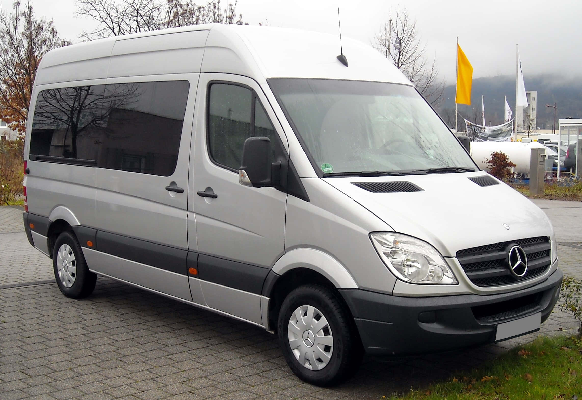 Mercedes Benz Sprinter 桌布 壁紙 — 一架時尚的銀色 Mercedes-Benz Sprinter 客貨車停泊在鋪砌的停車場，背景有光禿的樹木和霧氣籠罩的山脈。