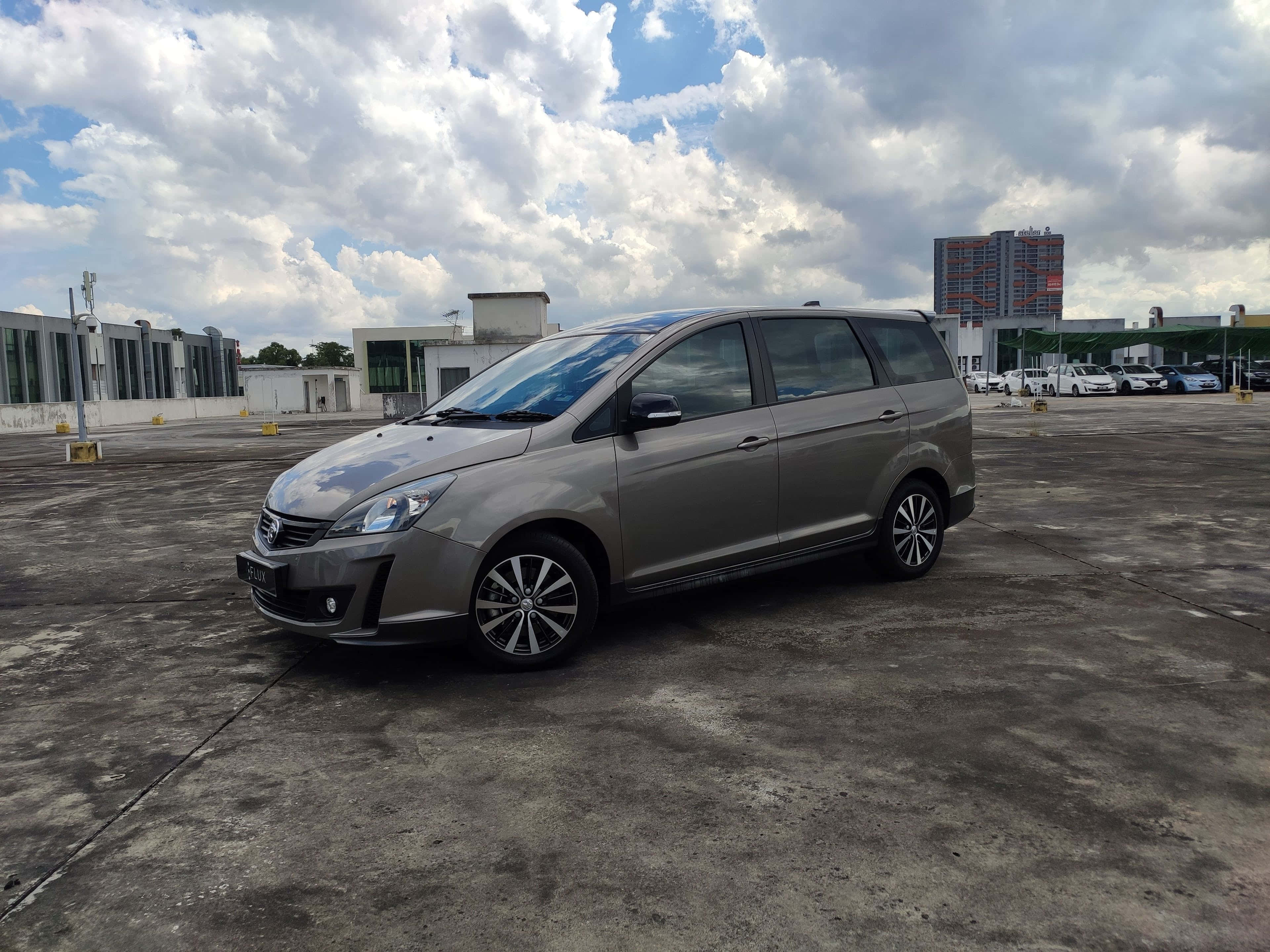Proton Exora 桌布 壁紙 — 一輛時尚的銀色小型貨車停泊在廣闊的混凝土場地上，背景是戲劇性的多雲天空，遠處有現代建築。