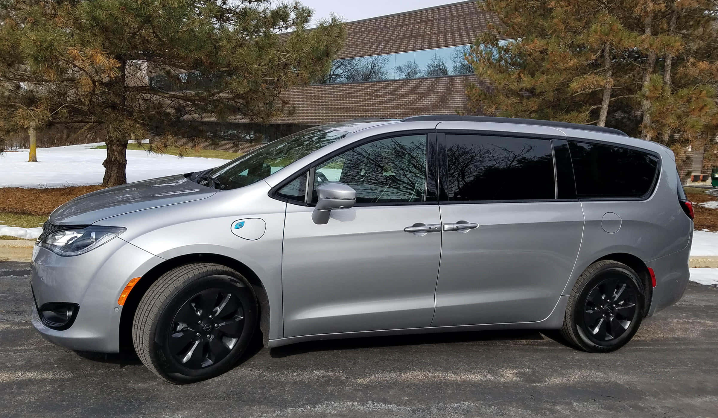 Chrysler Pacifica 桌布 壁紙 — 一輛時尚的銀色小型貨車配黑色輪圈，停泊在雪地車道上，旁邊有松樹和現代磚砌建築。