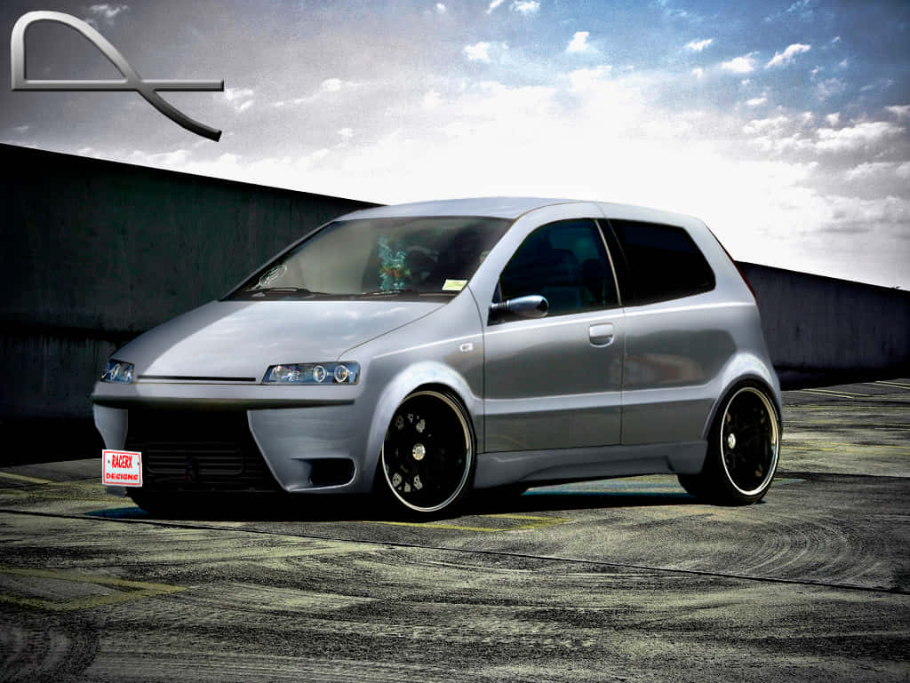 Fiat Grande Punto 桌布 壁紙 — 一輛時尚的銀色改裝掀背車，配有黑色輪圈，停在磨損的瀝青賽道上，天空多雲。