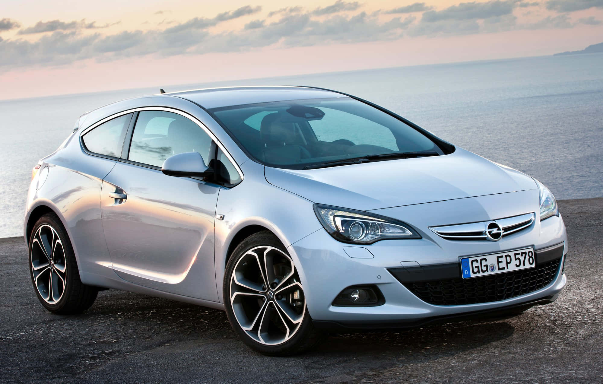 Opel Astra 桌布 壁紙 — 一輛時尚的銀色歐寶雅特雙門轎跑車停在日落時分的海岸公路上，背後是平靜的海水。