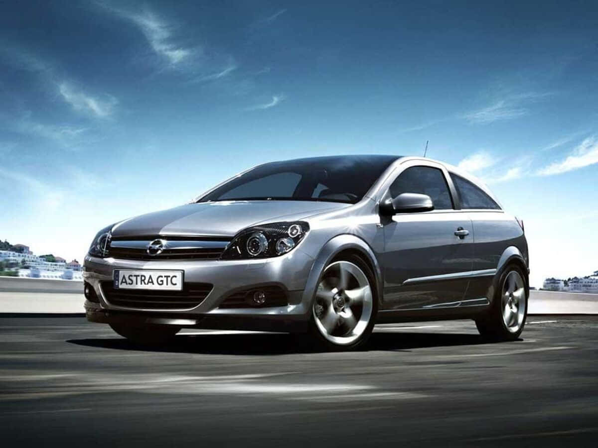 Opel Astra 桌布 壁紙 — 一輛時尚的銀色歐寶雅特GTC雙門轎跑車沿著海岸公路飛馳，天空湛藍，白雲輕飄。