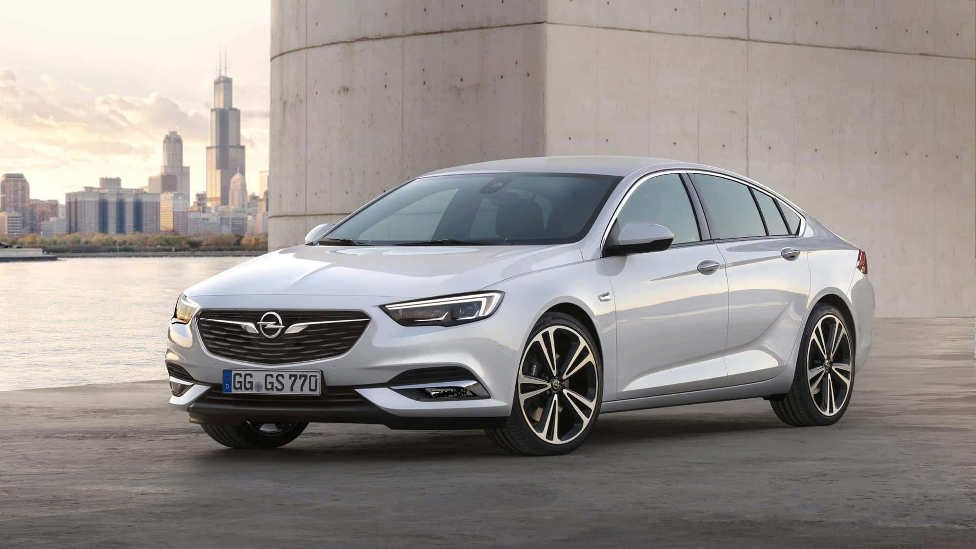 Opel Insignia 桌布 壁紙 — 一輛時尚的銀色歐寶Insignia GS停放在混凝土柱旁，背景是金色時刻的城市天際線和河流。