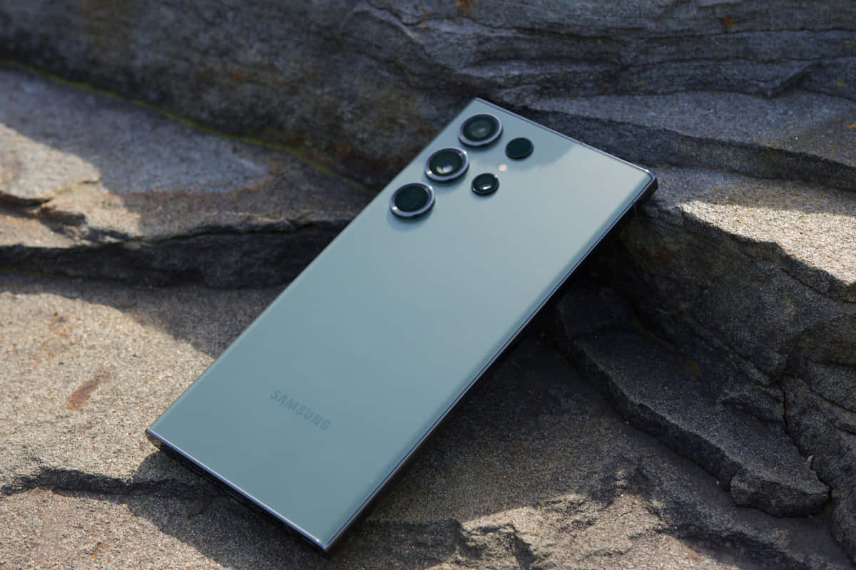 Samsung S23 Ultra 桌布 壁紙 — 一部時尚的銀色Samsung智能手機在明亮的陽光下斜放在有紋理的灰色岩石上。