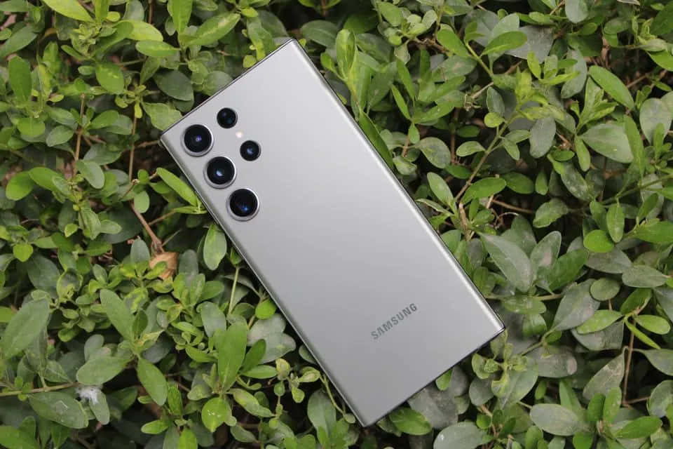 Samsung S23 Ultra 桌布 壁紙 — 一部時尚的銀色Samsung智能手機斜放在充滿活力的綠色葉子上，自然光突顯其相機陣列。