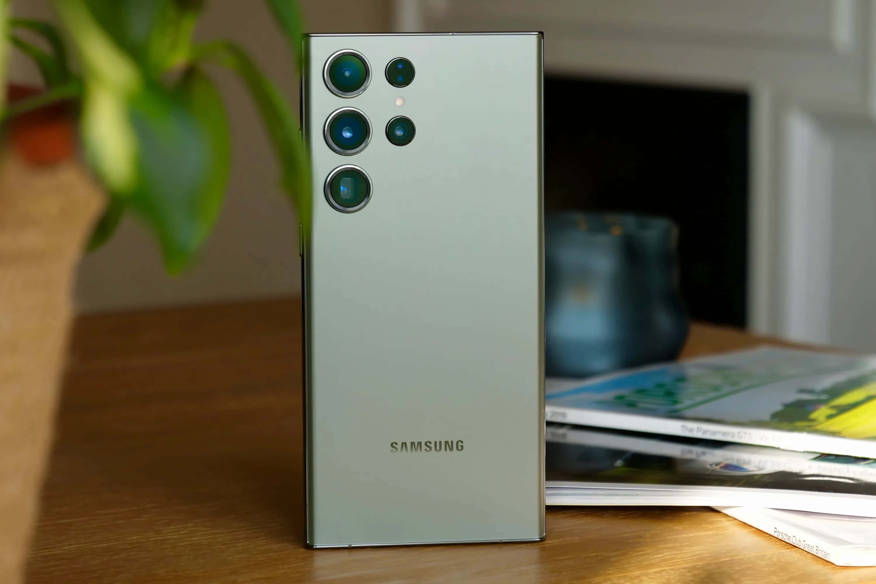 Samsung S23 Ultra 桌布 壁紙 — 一部時尚的銀色Samsung智能手機直立於木桌上，旁邊有雜誌和盆栽植物。