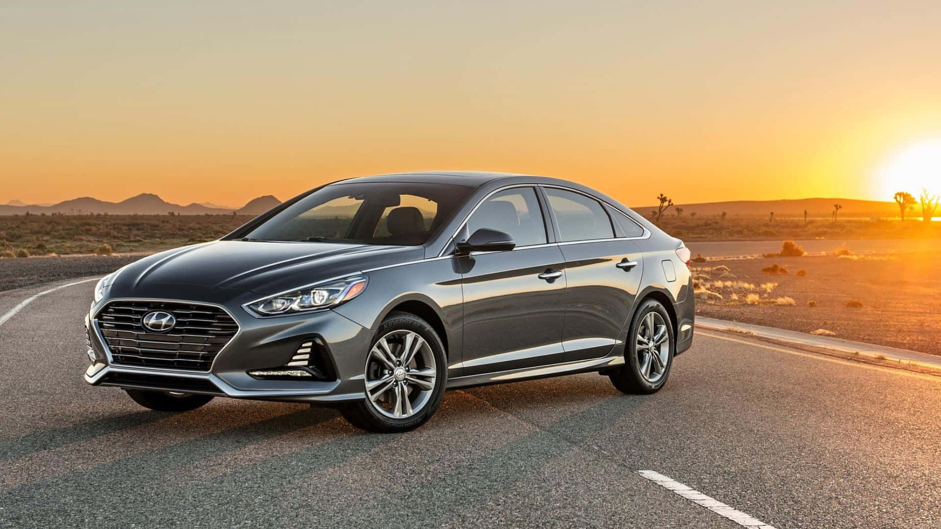 Hyundai Sonata 桌布 壁紙 — 一輛時尚的銀色轎車在沙漠公路上巡航，夕陽在遠山後落下，灑下金色光芒。