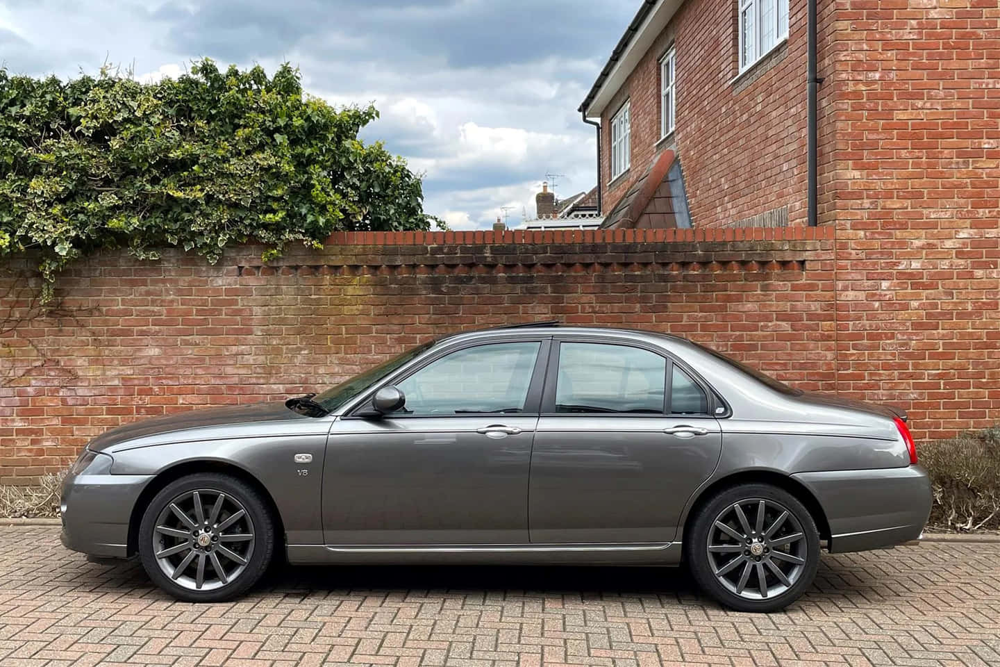 Mg Zt 桌布 壁紙 — 一架時尚的銀色房車停泊在磚砌車道上，旁邊是紅磚牆和茂密的樹籬，天空多雲。