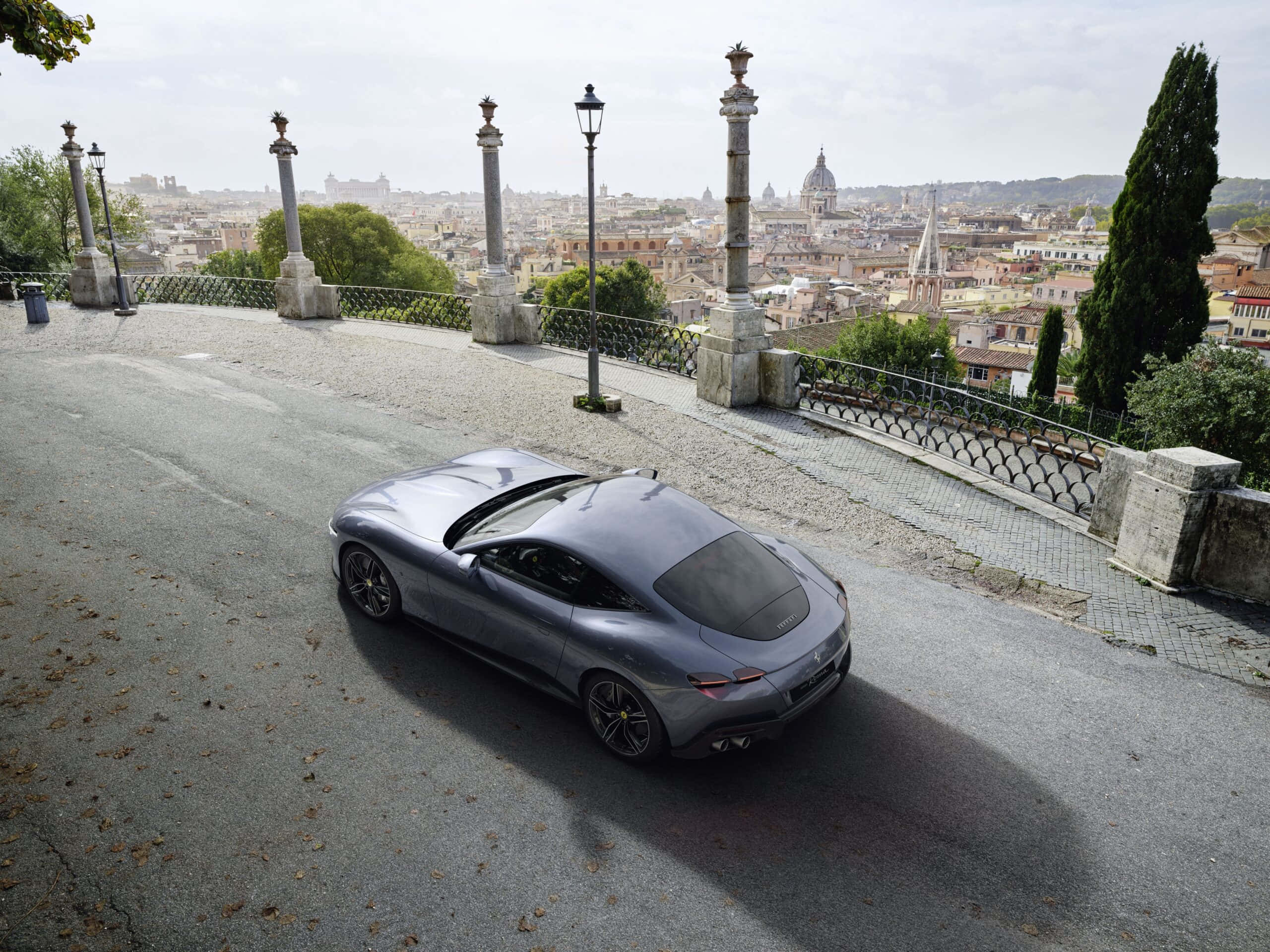 Ferrari Roma 桌布 壁紙 — 一架時尚的銀色跑車沿著鵝卵石山坡行駛，俯瞰著陰天下一片廣闊的歷史城市景觀。