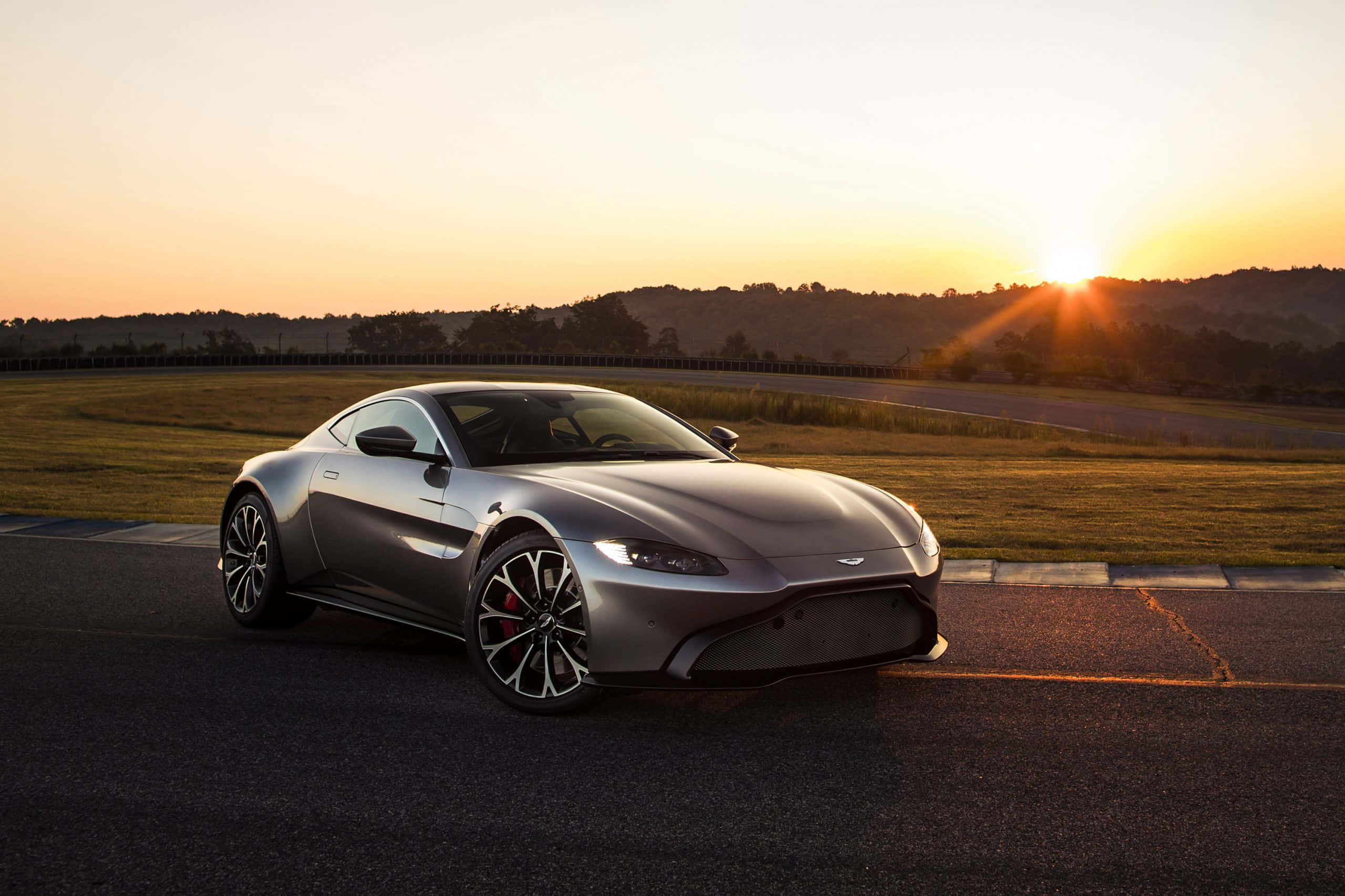 Aston Martin Vantage 桌布 壁紙 — 一輛流線型銀色跑車在賽道上飛馳，夕陽在起伏的山丘後落下，灑下金色光芒。