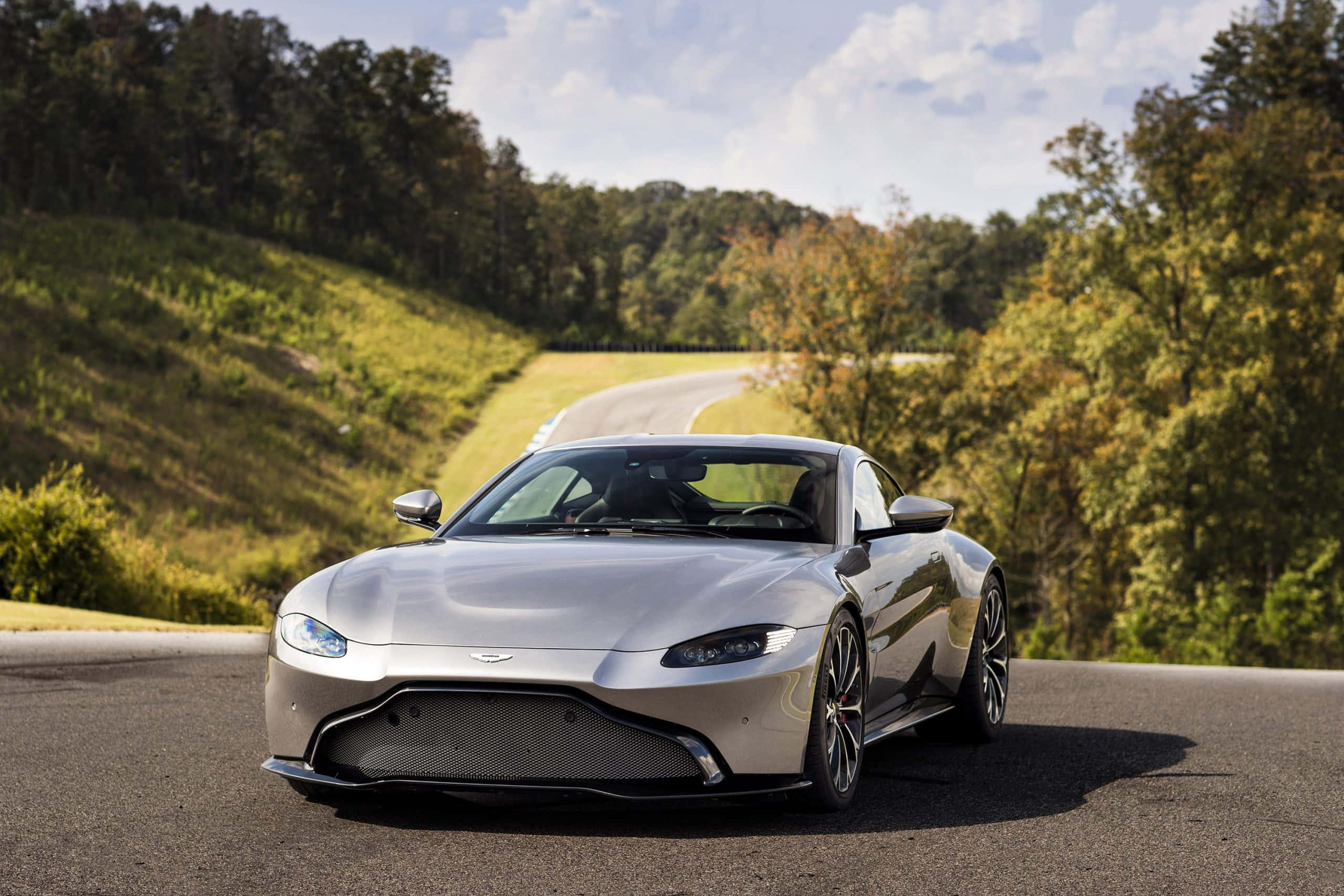 Aston Martin Vantage 桌布 壁紙 — 一輛時尚的銀色跑車沿著蜿蜒的森林道路行駛，在明亮的藍天下，有零星雲朵。