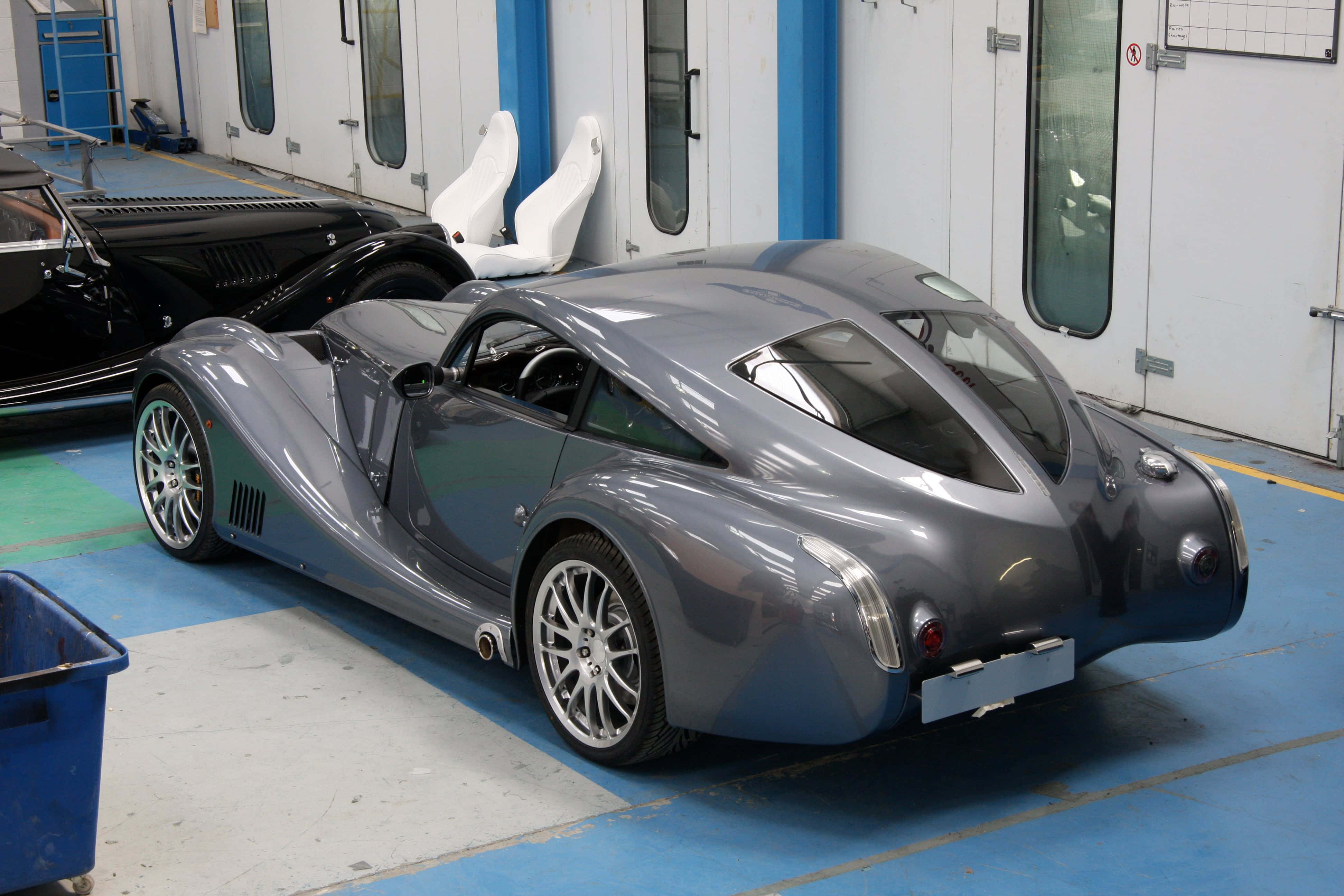 Morgan Aeromax 桌布 壁紙 — 一架流線型銀色跑車，帶有空氣動力學曲線，停放在乾淨的車庫中，旁邊有一輛黑色經典車和白色賽車座椅。
