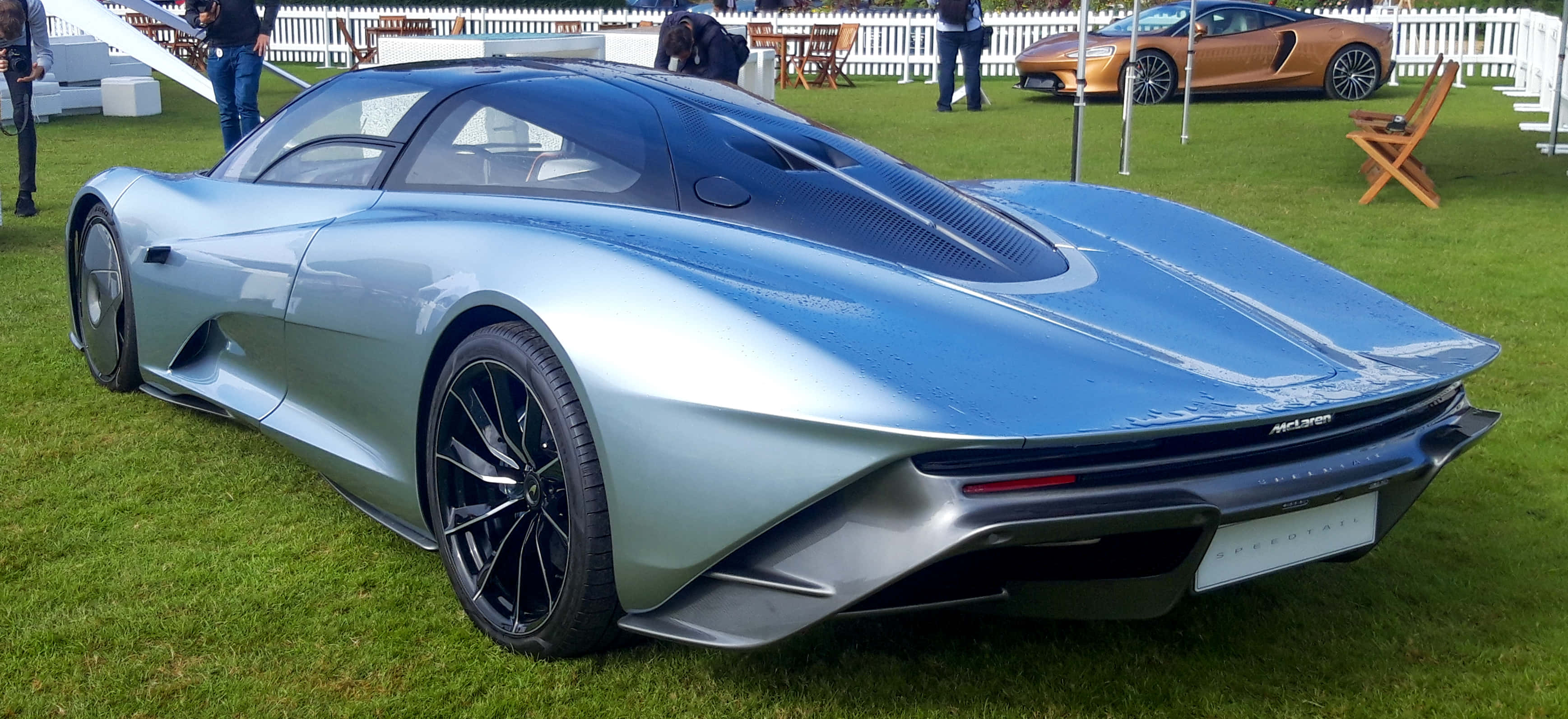 Mclaren Speedtail 桌布 壁紙 — 一輛時尚的銀色跑車帶有黑色飾邊，停在綠色草地上，背景是戶外車展，有人群和另一輛橙色汽車。