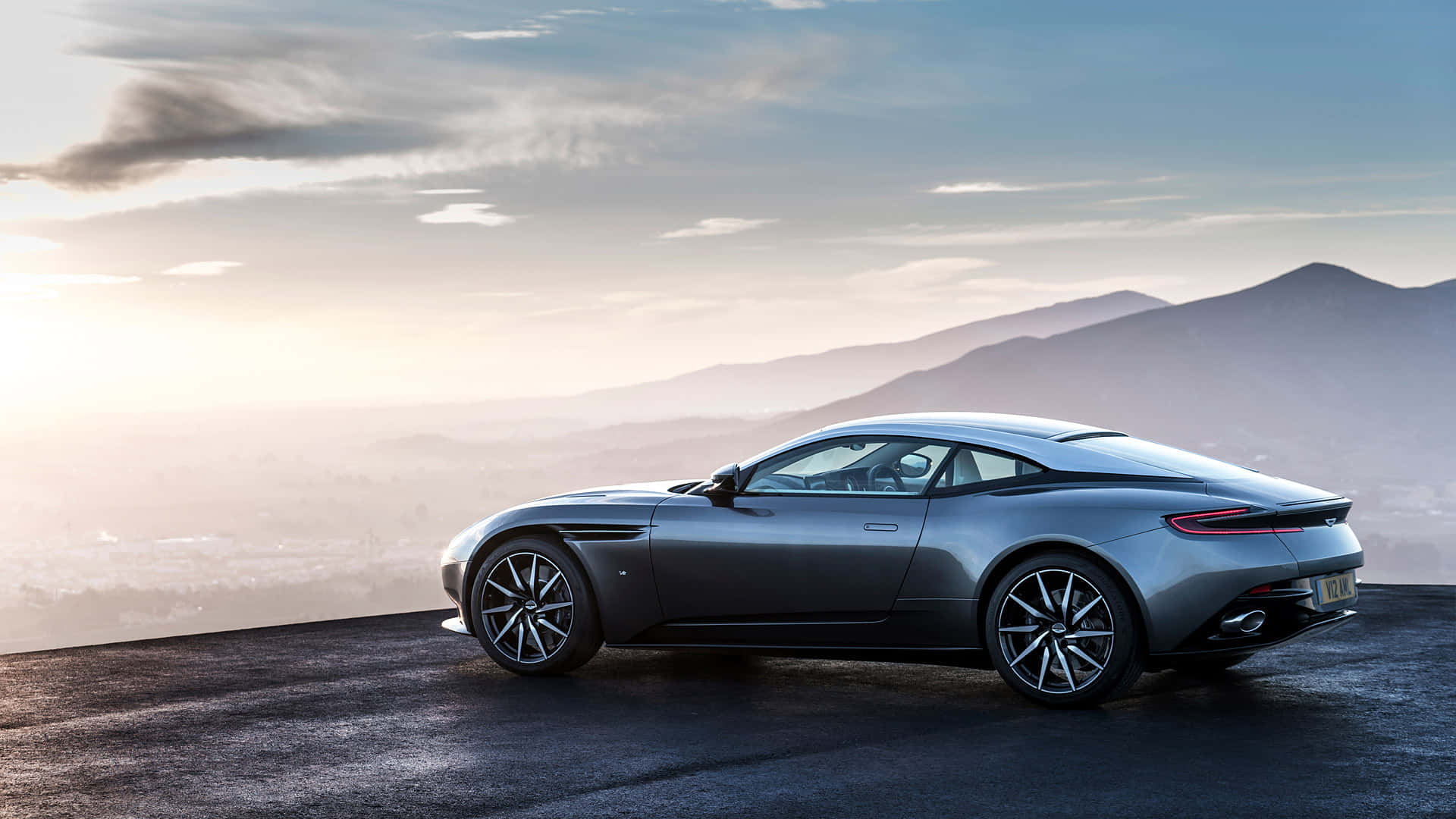 Aston Martin Db11 桌布 壁紙 — 一架時尚的銀色跑車停在懸崖上，俯瞰日出時遠處的山脈，伴隨著柔和的金色光芒。