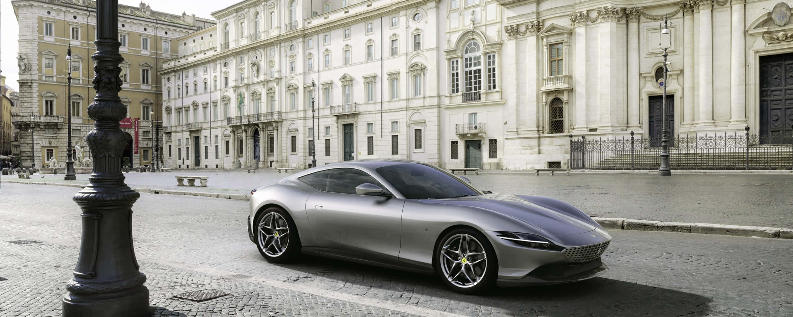 Ferrari Roma 桌布 壁紙 — 一輛時尚的銀色跑車在明亮的日光下停泊在鵝卵石街道上，前方是宏偉的古典建築。