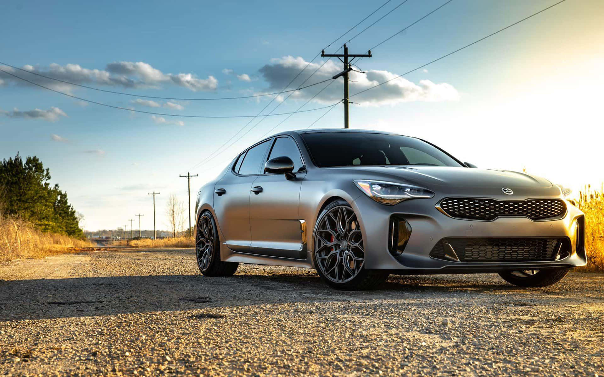 Kia Stinger 桌布 壁紙 — 一架時尚的銀色跑車停泊在碎石路上，上方是晴朗藍天，點綴著幾朵白雲。