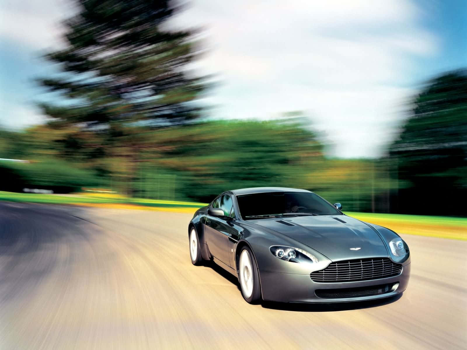 Aston Martin Vantage 桌布 壁紙 — 一輛時尚的銀色跑車在蜿蜒的道路上飛馳，背景是模糊的綠樹和天空。