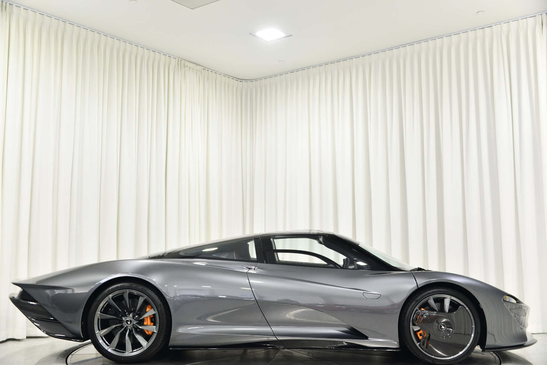 Mclaren Speedtail 桌布 壁紙 — 一輛時尚的銀色跑車配備橙色制動卡鉗，在極簡主義工作室中以白色窗簾為背景展示。