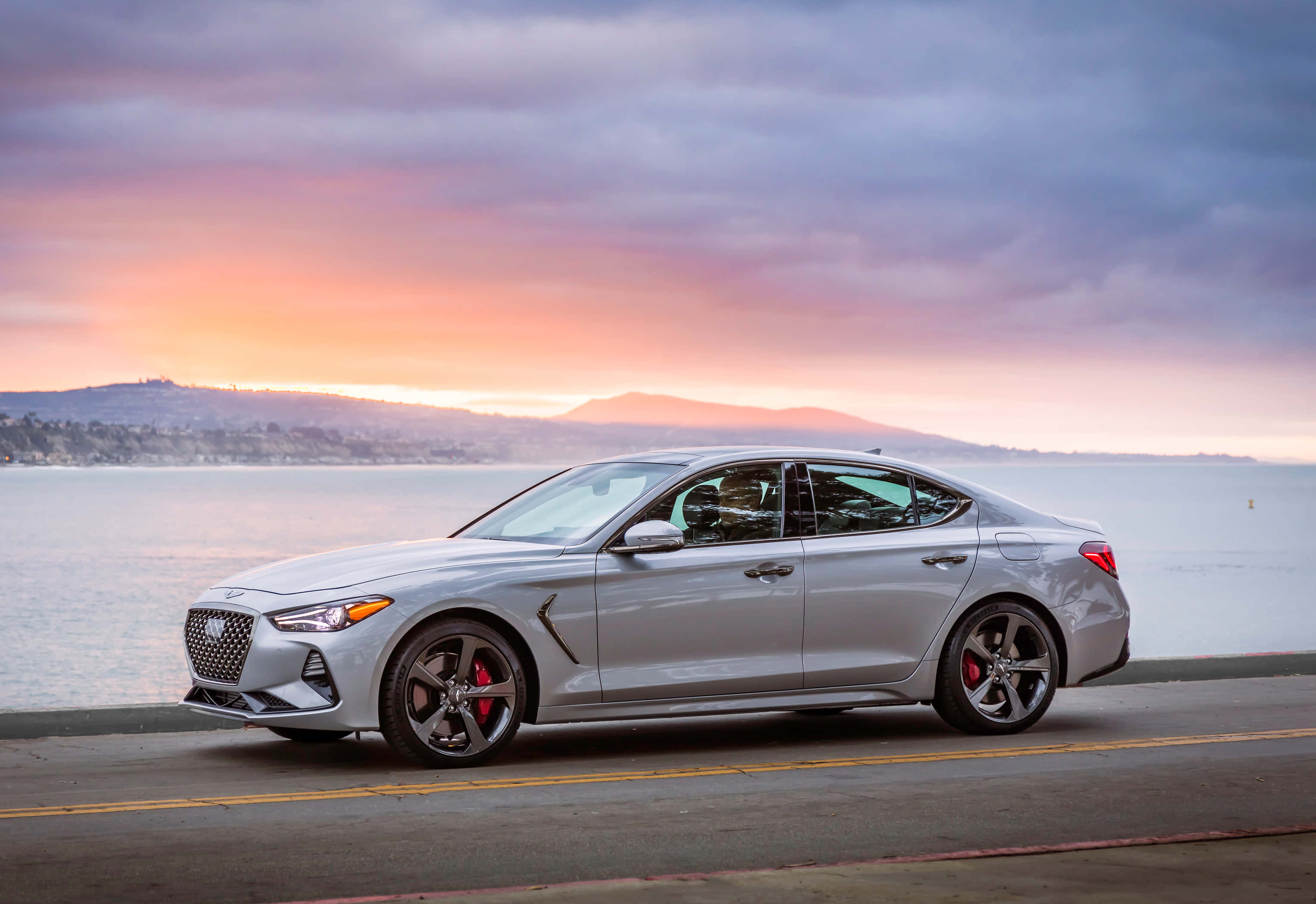 Genesis G70 桌布 壁紙 — 一輛時尚的銀色跑車配備紅色制動卡鉗，停在日落時分的海濱道路上，天空呈現粉紅色和紫色。