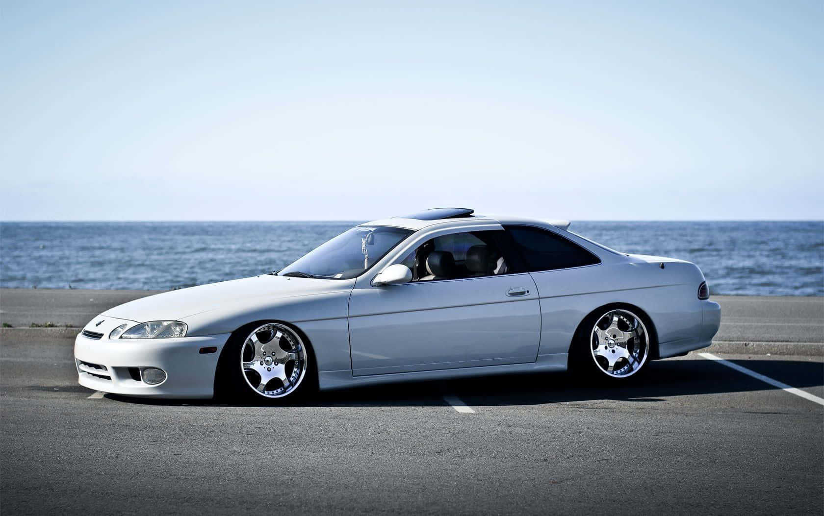 Lexus Sc 300 桌布 壁紙 — 一輛時尚的銀色運動轎跑車配備定制輪圈，停在沿海公路上，旁邊是海洋，天空清澈湛藍。