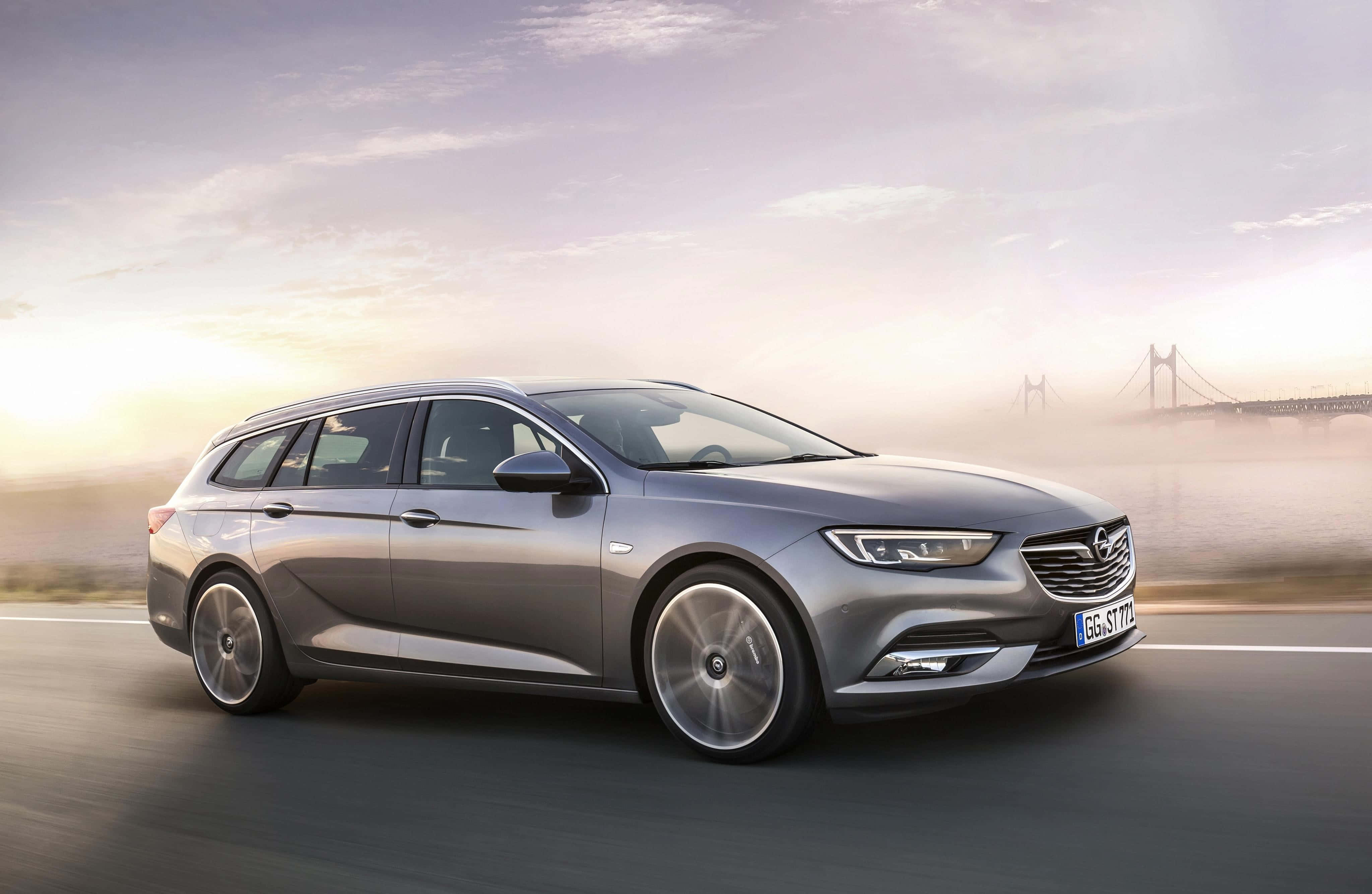 Opel Insignia 桌布 壁紙 — 一架時尚的銀色旅行車沿著海岸公路在日出時行駛，遠處有一座霧濛濛的橋樑。