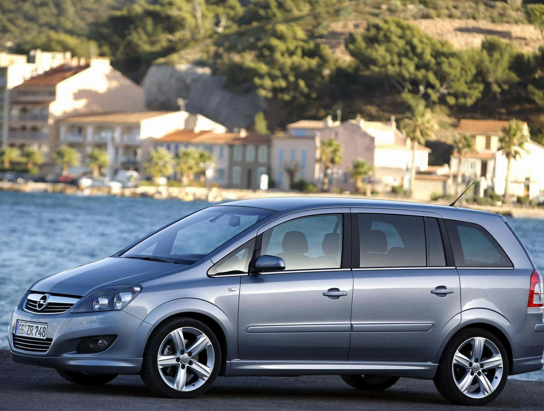 Opel Zafira 桌布 壁紙 — 一架時尚的銀色旅行車停在海濱，背景是地中海風格的房屋和綠色山丘。