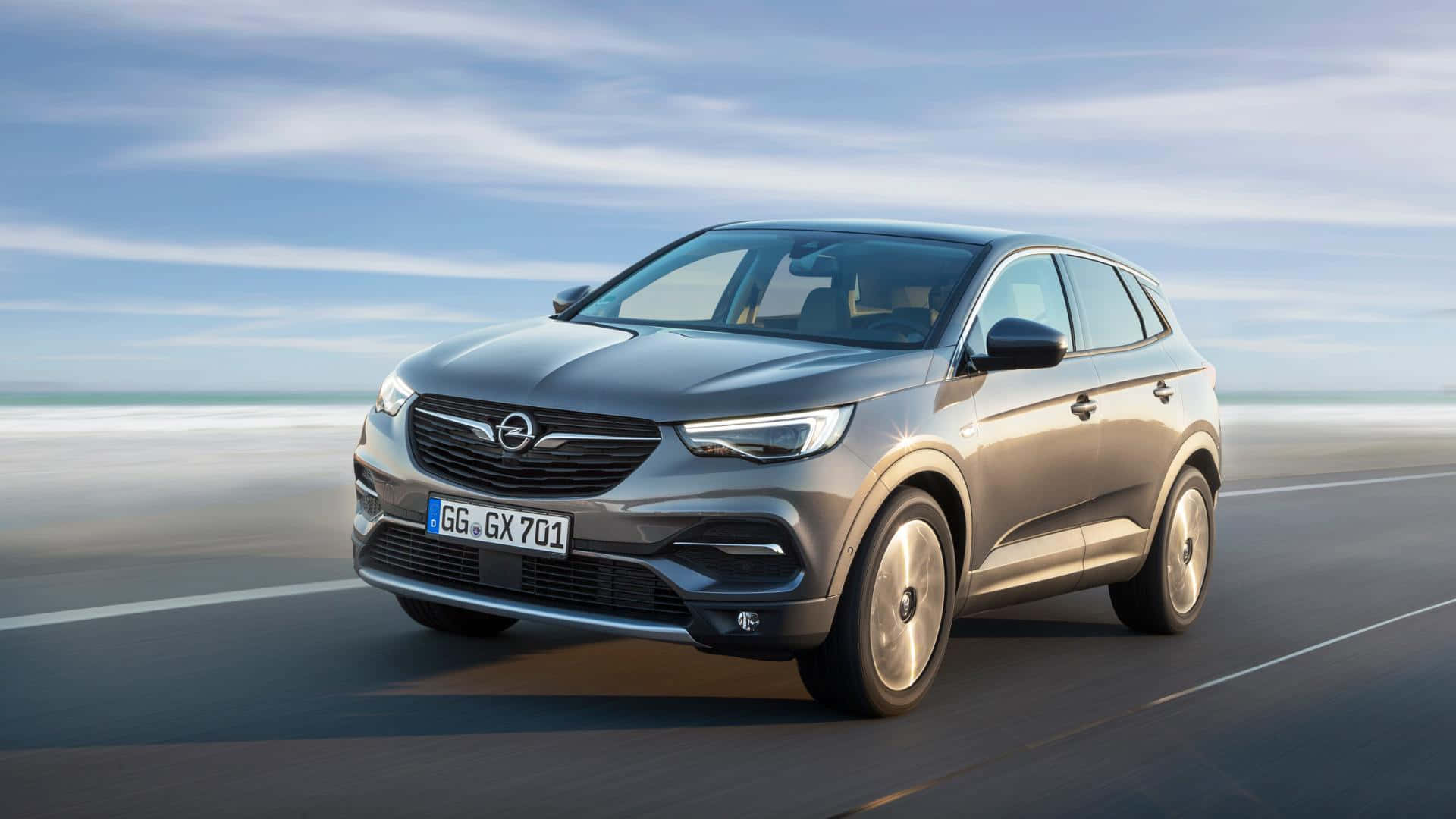 Opel Grandland X 桌布 壁紙 — 一輛時尚的銀色SUV沿著海岸道路行駛，背景是模糊的海洋和天空。