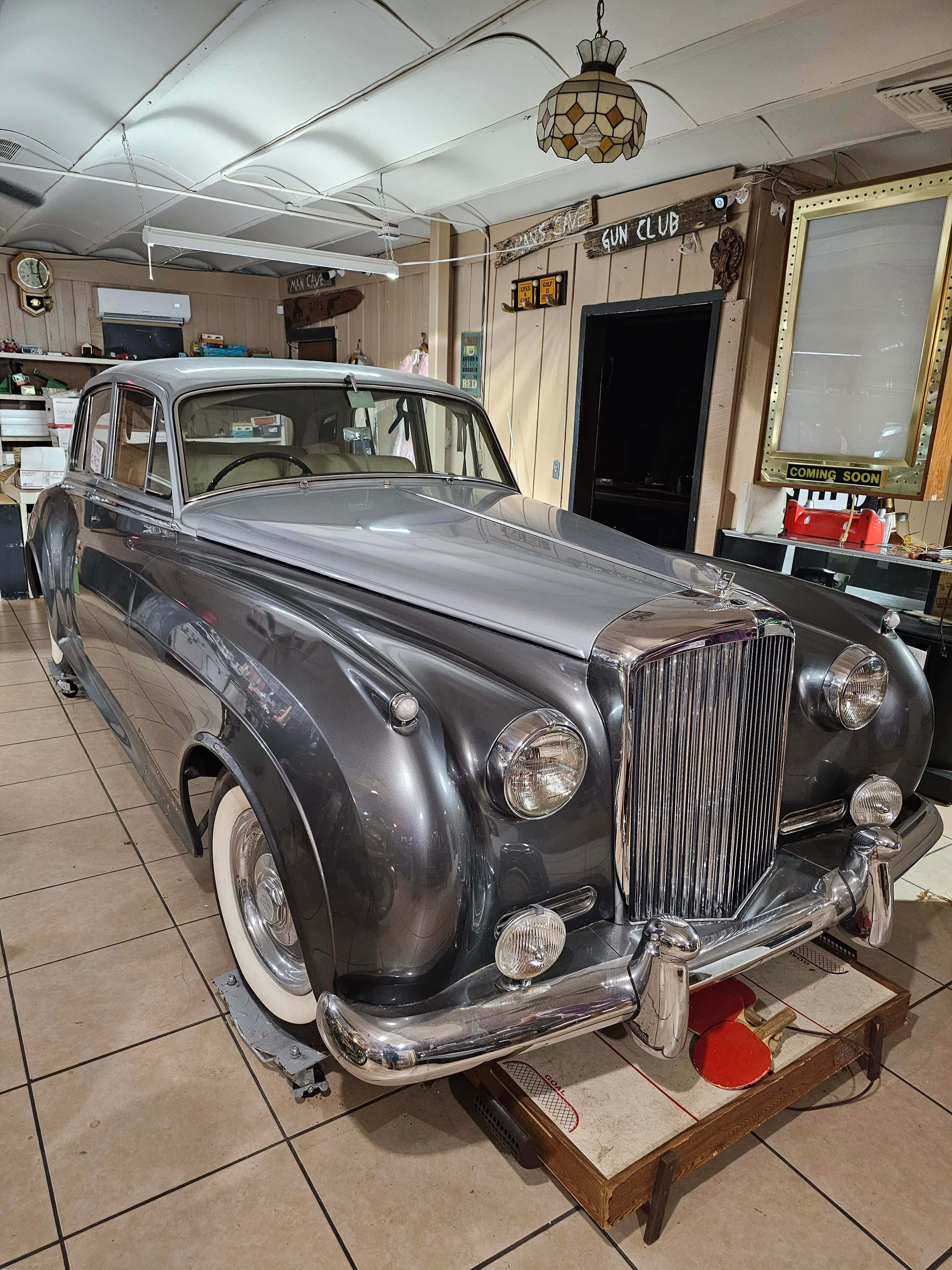 Bentley S2 桌布 壁紙 — 一輛時尚的銀色復古車在舖有瓷磚的車庫中，在溫暖的燈光下閃閃發光，車庫裝飾著復古標誌和裝飾品。