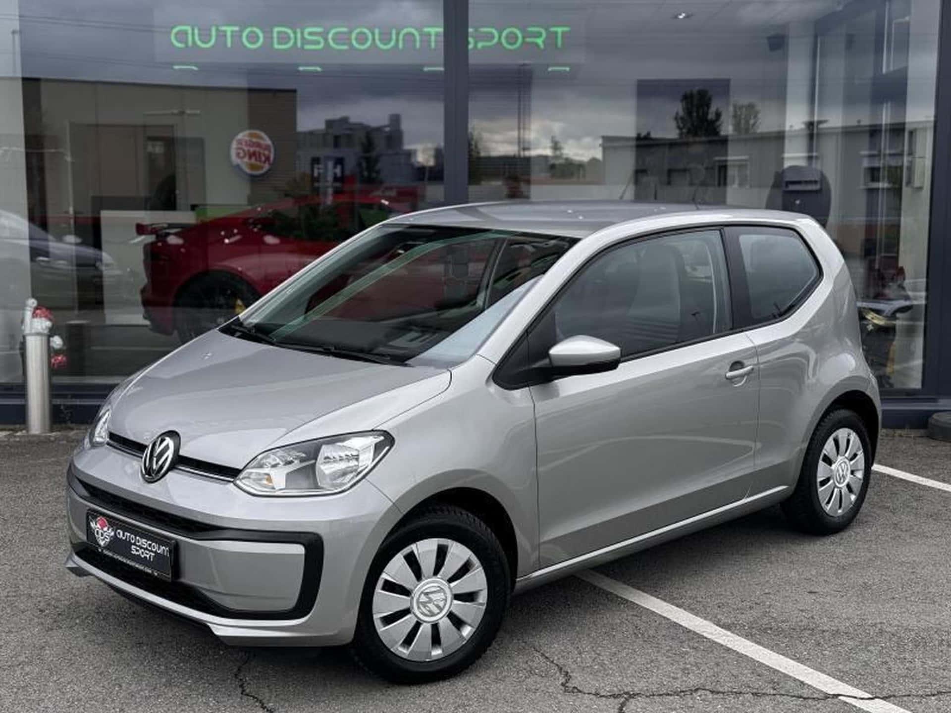  壁紙 — 一輛時尚的銀色Volkswagen up!停泊在玻璃窗反射多雲天空和內部紅色汽車的汽車經銷店前。