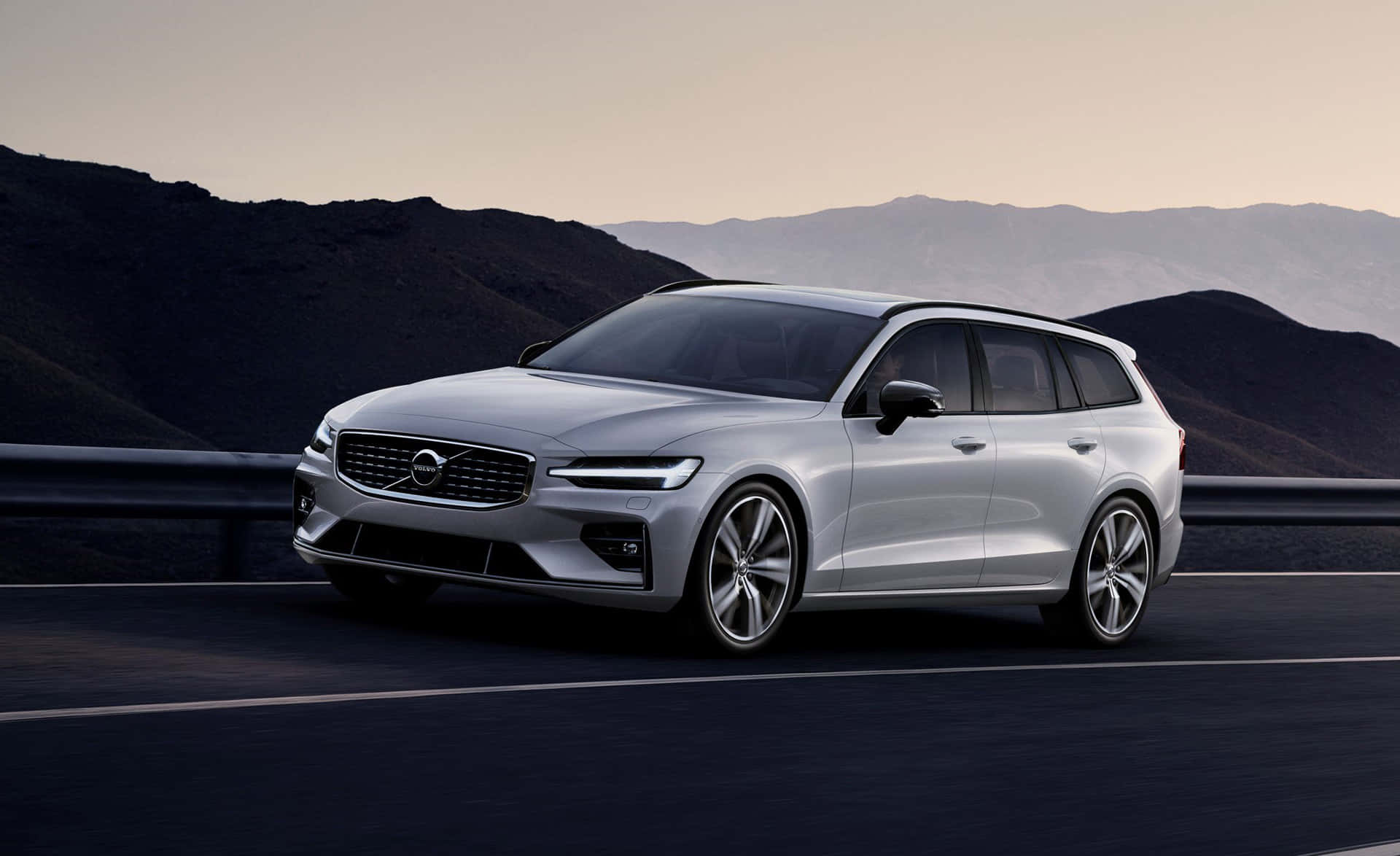 Volvo V60 桌布 壁紙 — 一架時尚的銀色富豪旅行車在黃昏時分沿著山區高速公路行駛，環境燈光柔和。