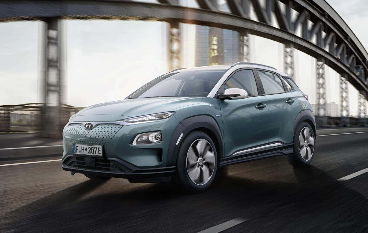 Hyundai Kona 桌布 壁紙 — 一輛時尚的藍綠色電動SUV在鋼拱橋下行駛，道路上有動態模糊效果。