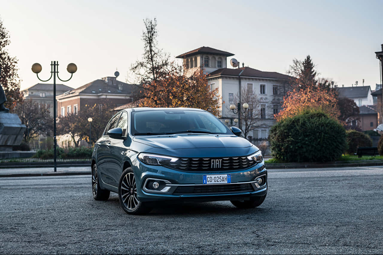 Fiat Tipo 桌布 壁紙 — 一架時尚的藍綠色快意轎車停泊在鵝卵石廣場上，周圍是秋樹和歷史建築，在柔和日光下。