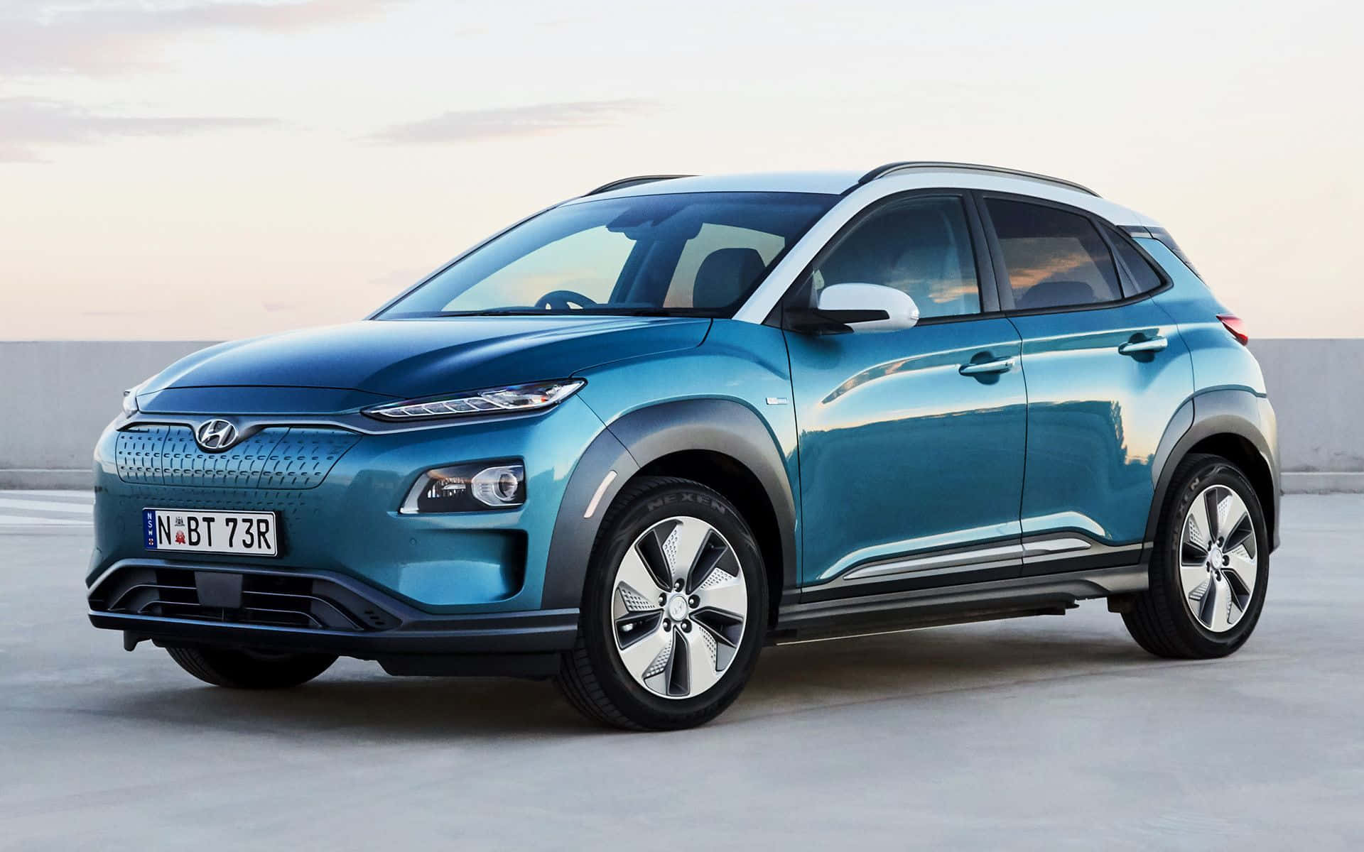 Hyundai Kona 桌布 壁紙 — 一架時尚嘅青綠色Hyundai Kona電動SUV停泊喺混凝土表面，喺柔和嘅暮色天空下。