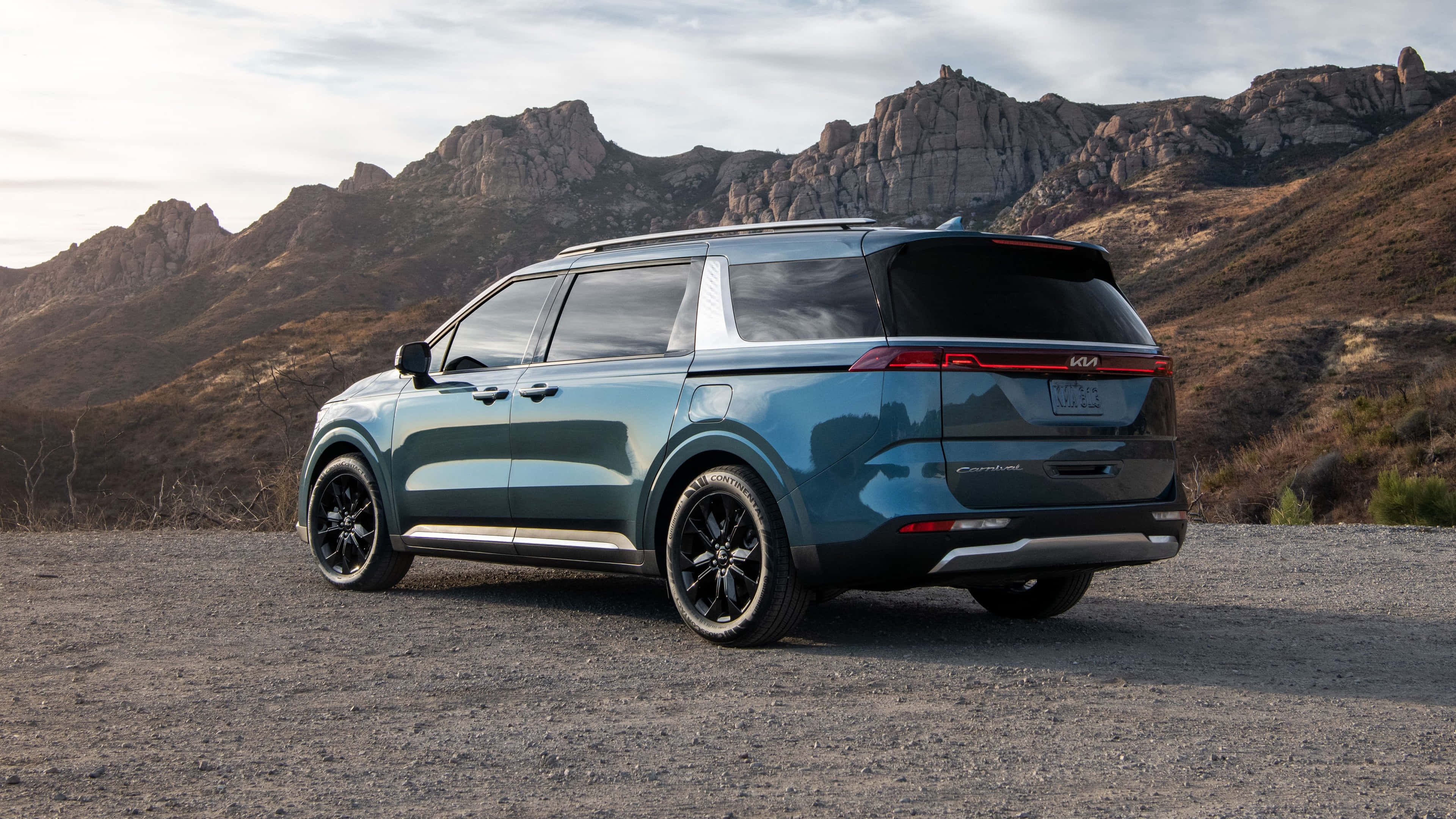Kia Carnival 桌布 壁紙 — 一架時尚的青色Kia小型貨車停在碎石上，背景是崎嶇的山脈，天空柔和。