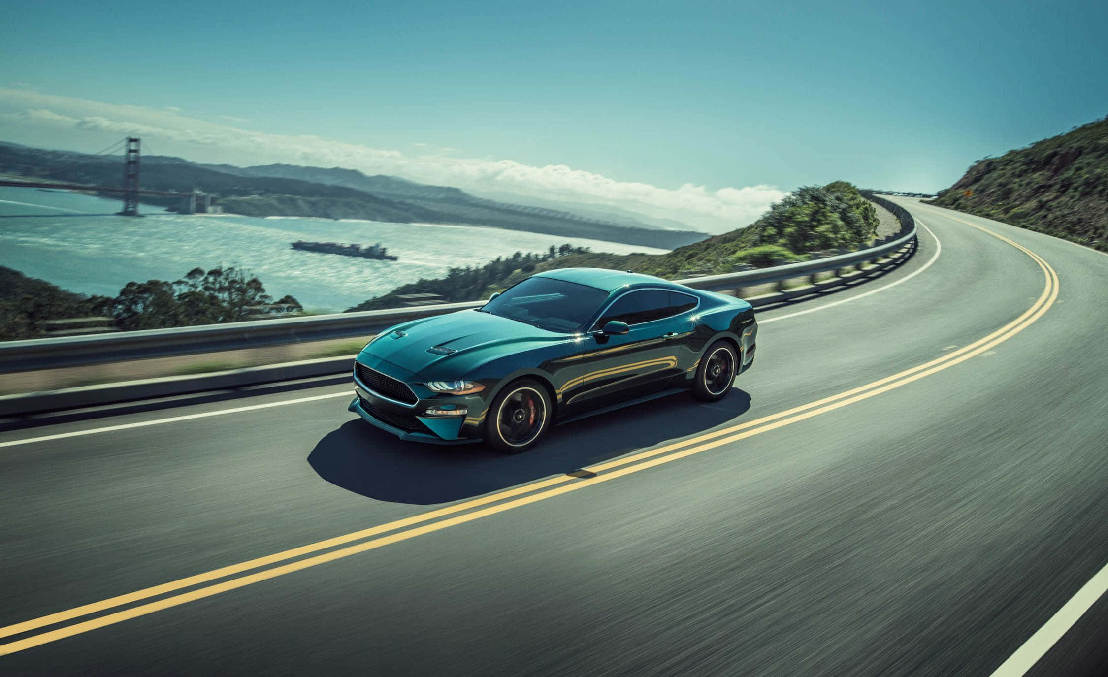 Ford Mustang Bullitt 桌布 壁紙 — 一架時尚的青綠色Mustang沿著海岸高速公路飛馳，遠處的金門大橋在晴朗的藍天下清晰可見。
