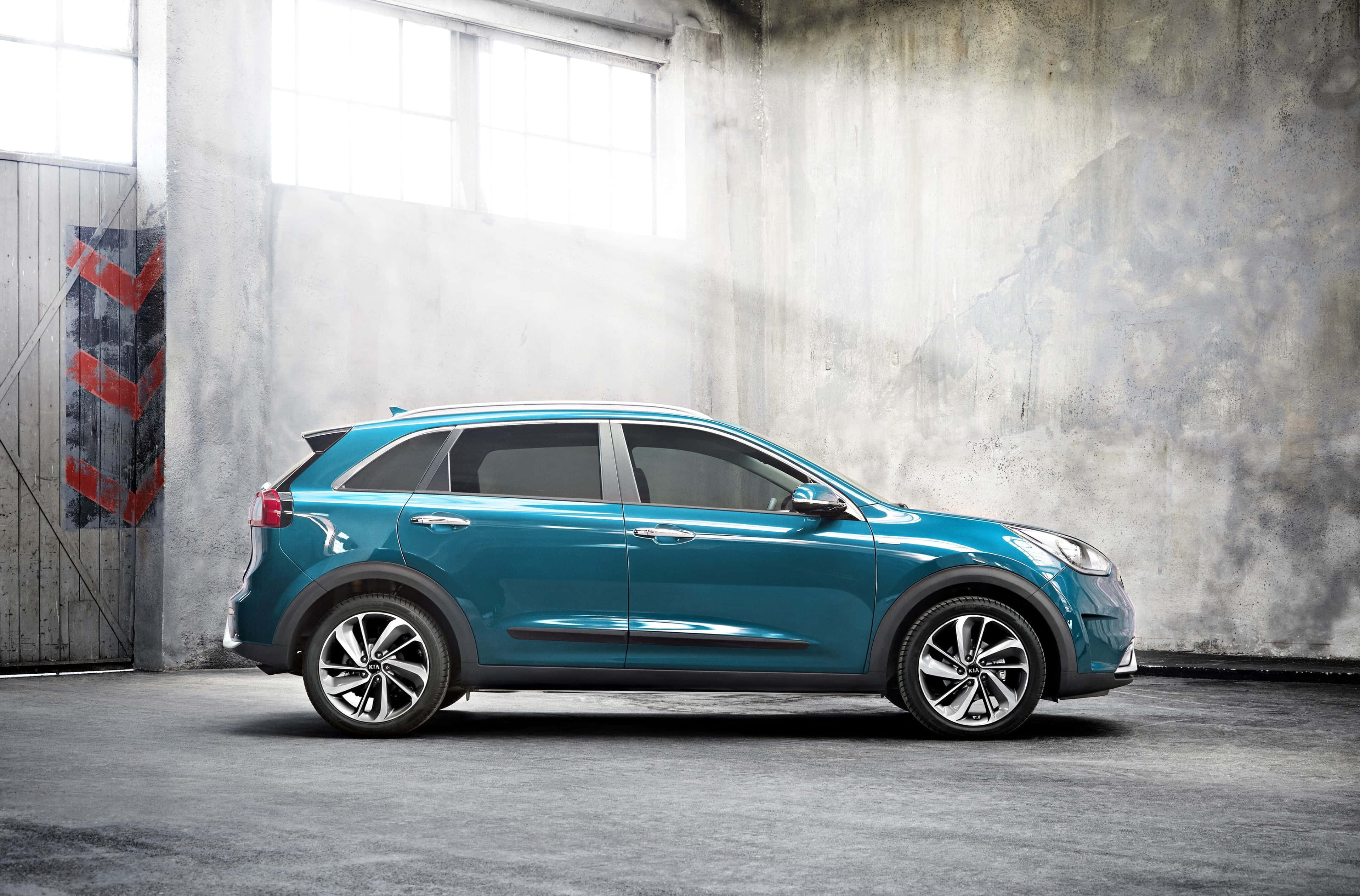 Kia Niro 桌布 壁紙 — 一架時尚的青綠色SUV側面停放在一個粗糙的混凝土倉庫內，灰塵彌漫的光線從高窗射入。