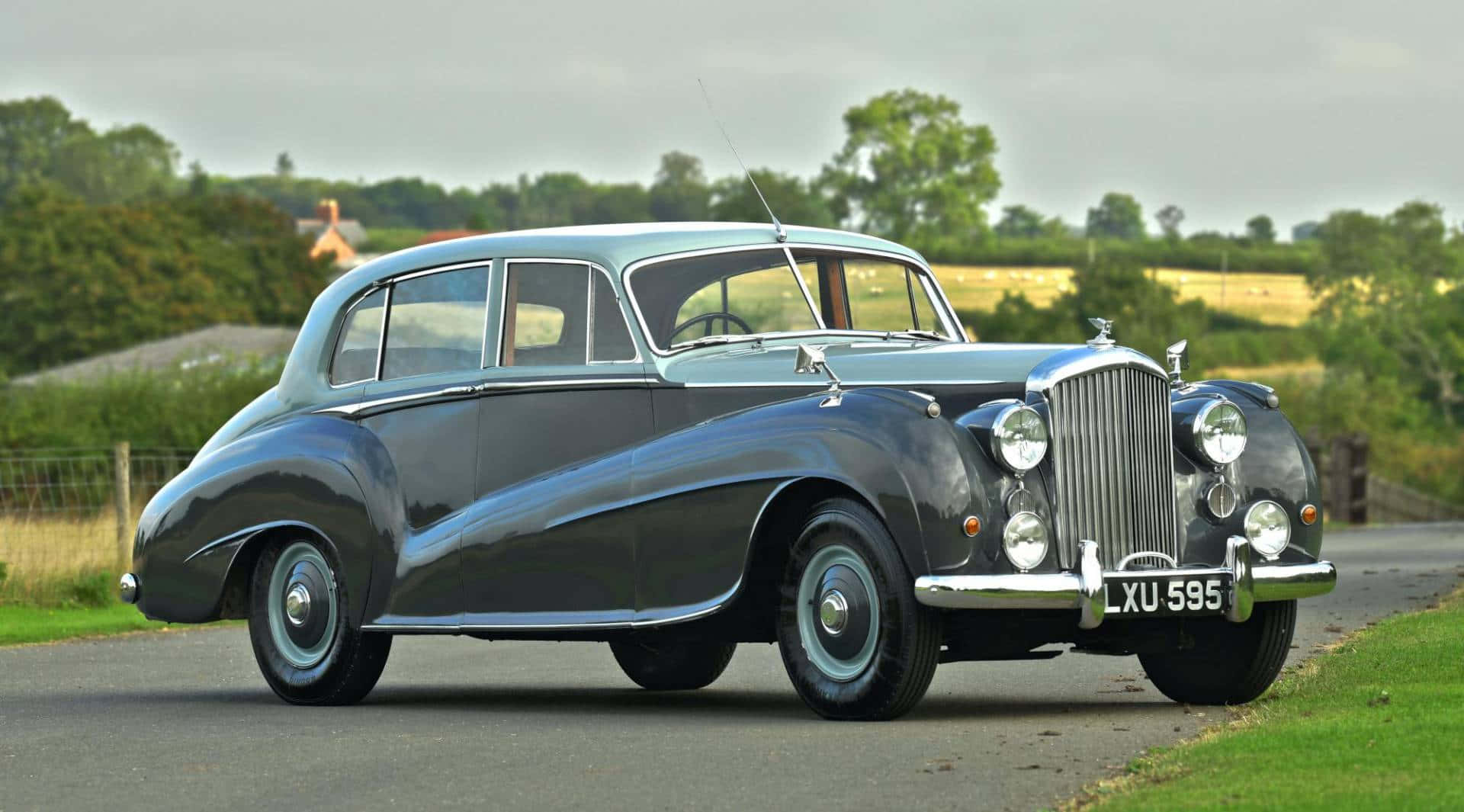 Bentley Mark Vi 桌布 壁紙 — 一架時尚的復古賓利房車在陰天之下閃閃發光，停泊在鄉村道路旁，旁邊是茂密的綠色田野和樹籬。