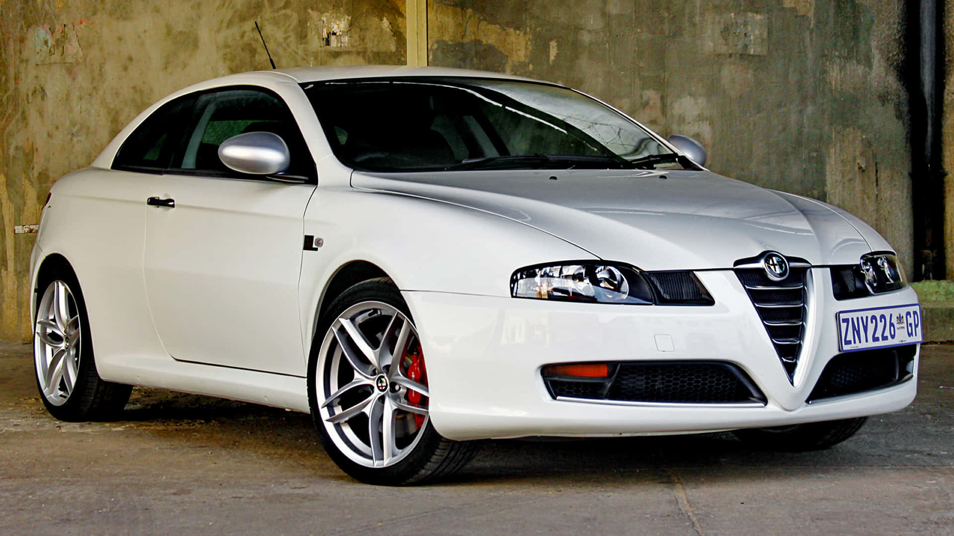 Alfa Romeo Gt 桌布 壁紙 — 一輛時尚的白色愛快羅密歐雙門跑車，帶有紅色制動卡鉗，停放在風化的混凝土牆旁。