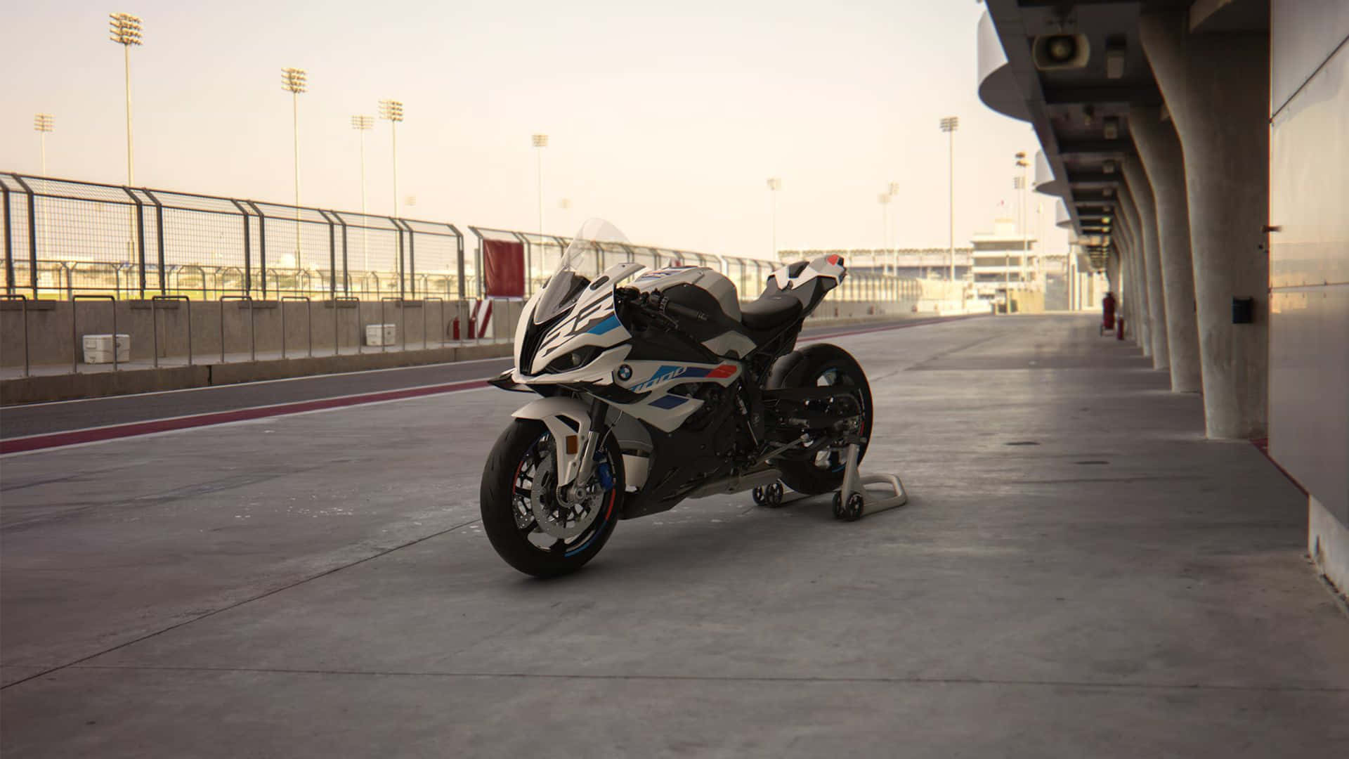  壁紙 — 一架時尚的白色和藍色BMW運動電單車，在溫暖的黃金時段光線下，停泊在賽車場的混凝土維修區車道上。