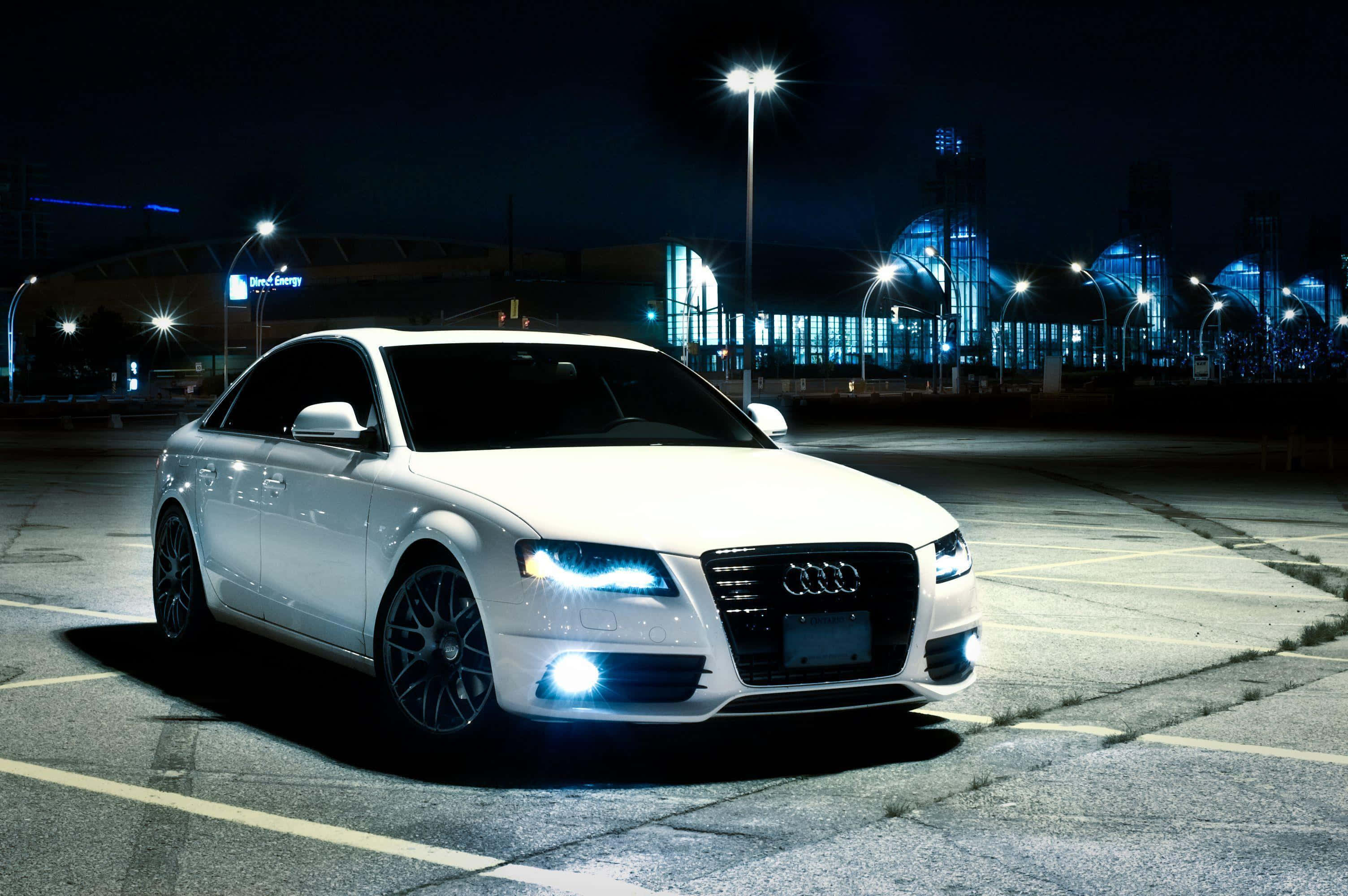 Audi A4 桌布 壁紙 — 一輛時尚的白色奧迪轎車在明亮的車頭燈下閃閃發光，背景是夜晚空蕩蕩的停車場和閃爍的城市燈光。
