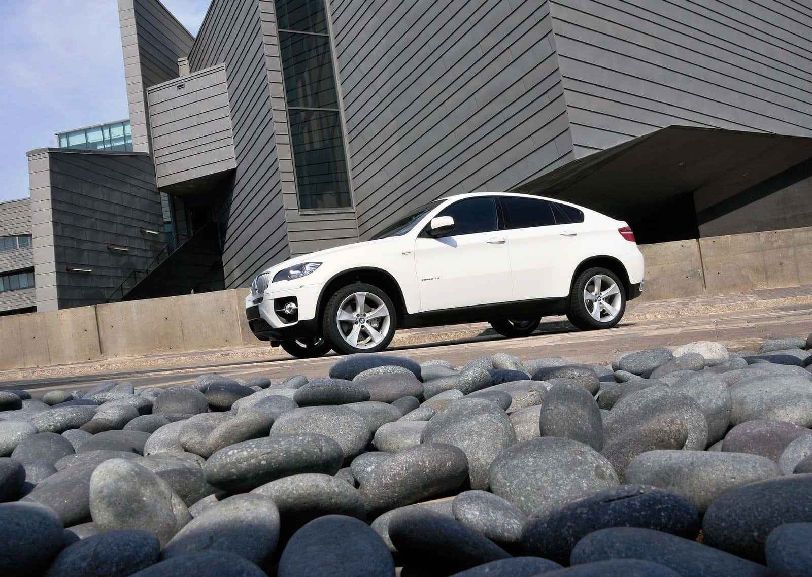 Bmw X6 桌布 壁紙 — 一架時尚的白色BMW X6 SUV停泊在現代灰色建築物旁邊的道路上，前景是光滑的灰色河石。