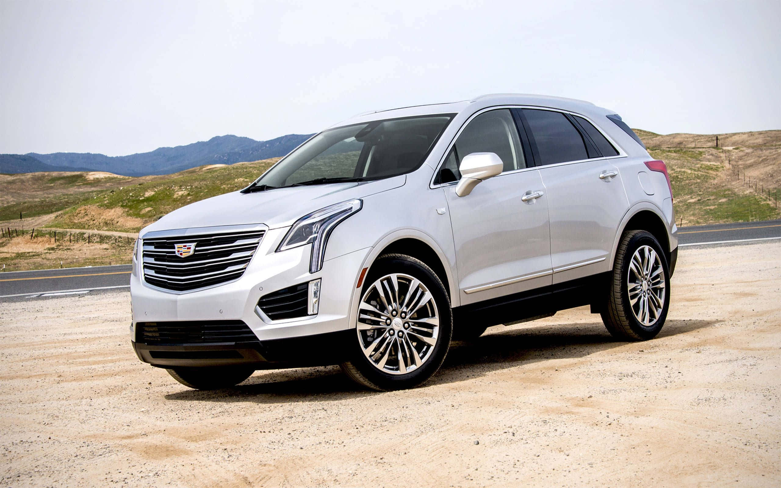 Cadillac Xt5 桌布 壁紙 — 一輛時尚的白色凱迪拉克SUV停在塵土飛揚的路邊，背景是起伏的丘陵和遠處的山脈，天空明亮。