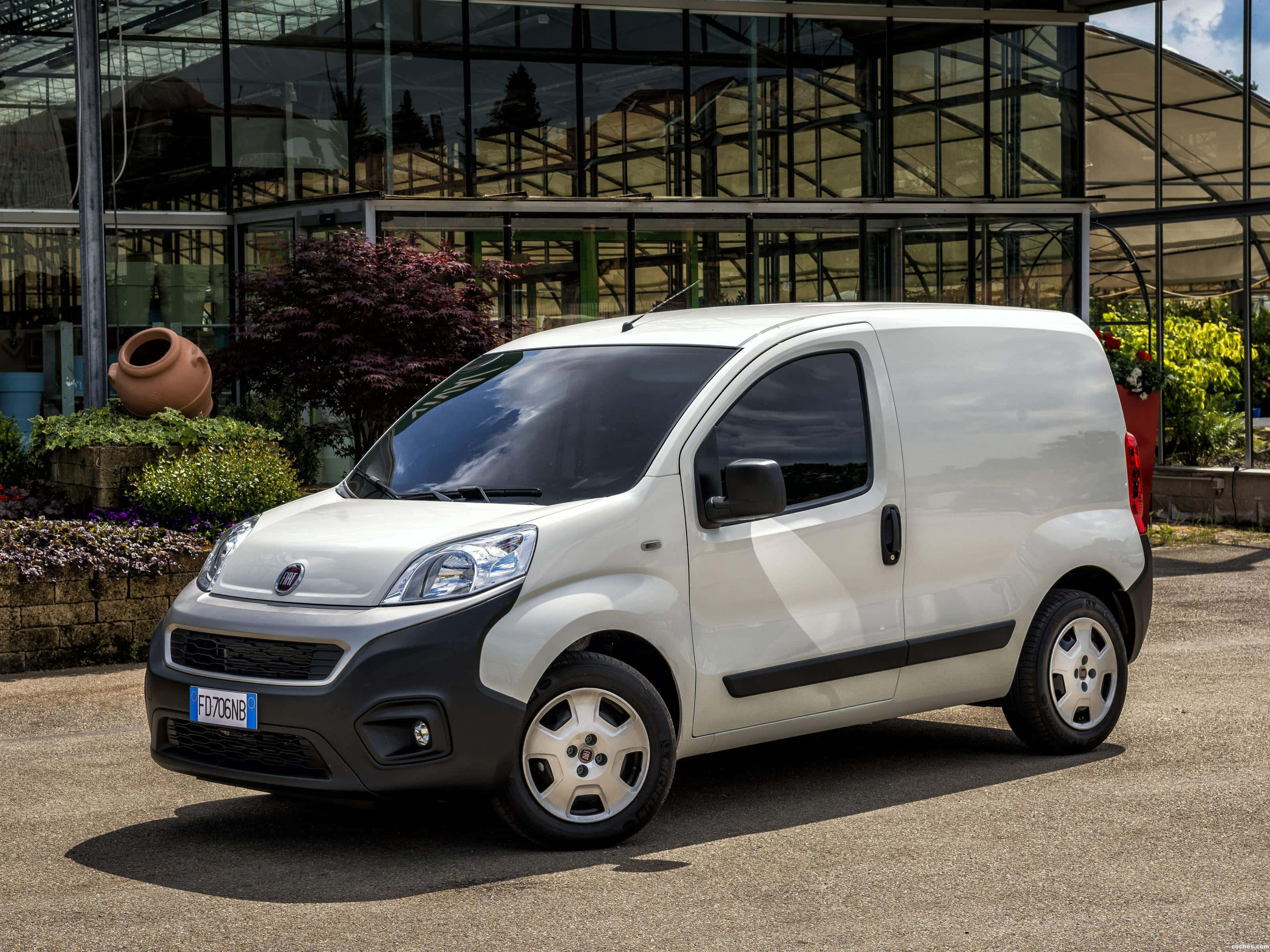 Fiat Fiorino 桌布 壁紙 — 一架時尚的白色 Fiat Fiorino 客貨車停泊在碎石上，前面是現代玻璃溫室，周圍綠意盎然。