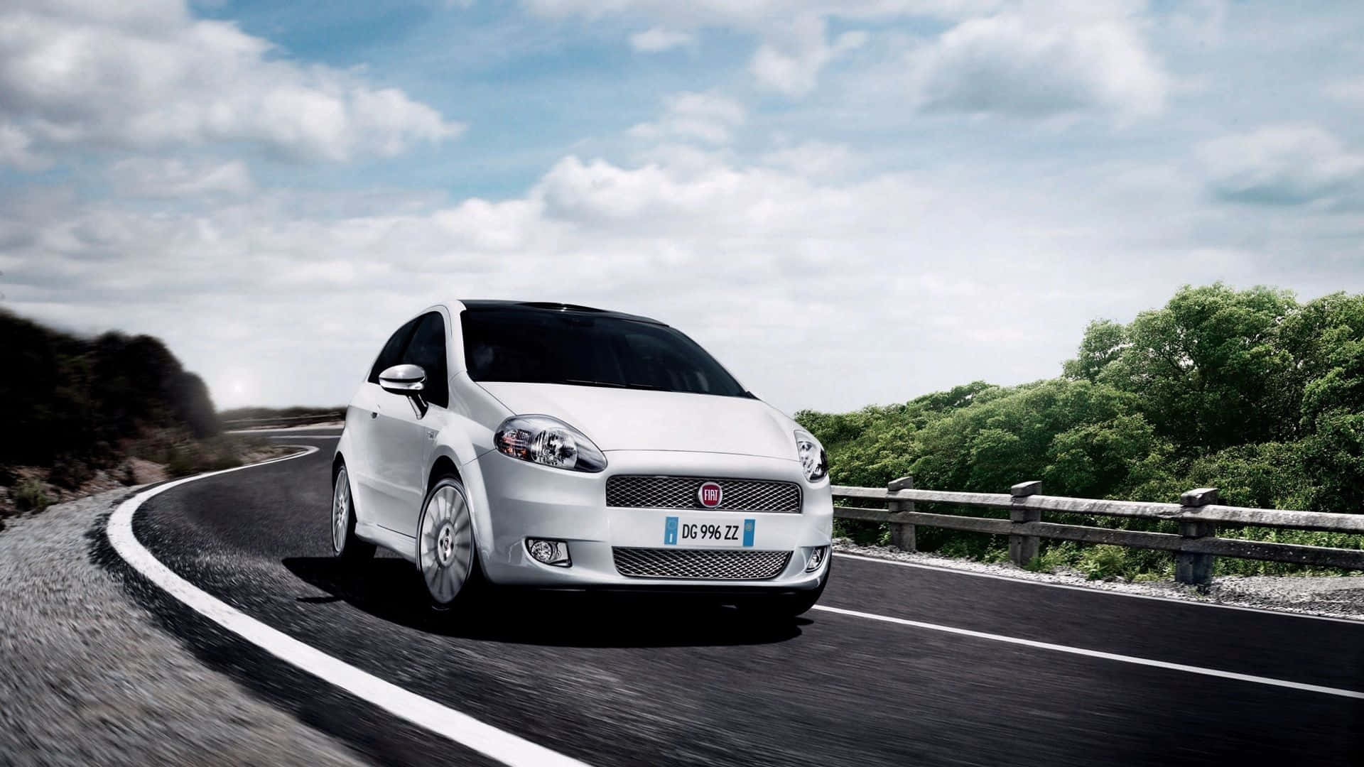 Fiat 500l 桌布 壁紙 — 一輛時尚的白色 Fiat Punto 沿著蜿蜒的瀝青道路行駛，兩旁綠樹成蔭，天空部分多雲。