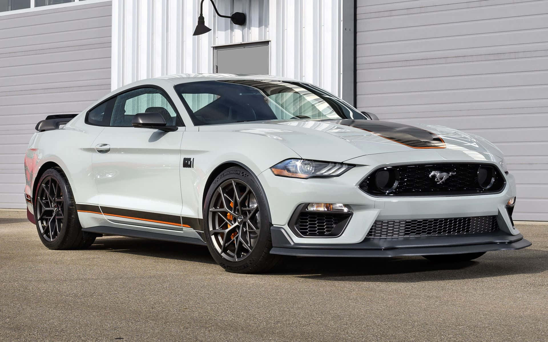 Ford Mustang Mach 1 桌布 壁紙 — 一輛時尚的白色福特野馬，帶有黑色點綴和橙色飾條，停在一棟現代工業建築前。