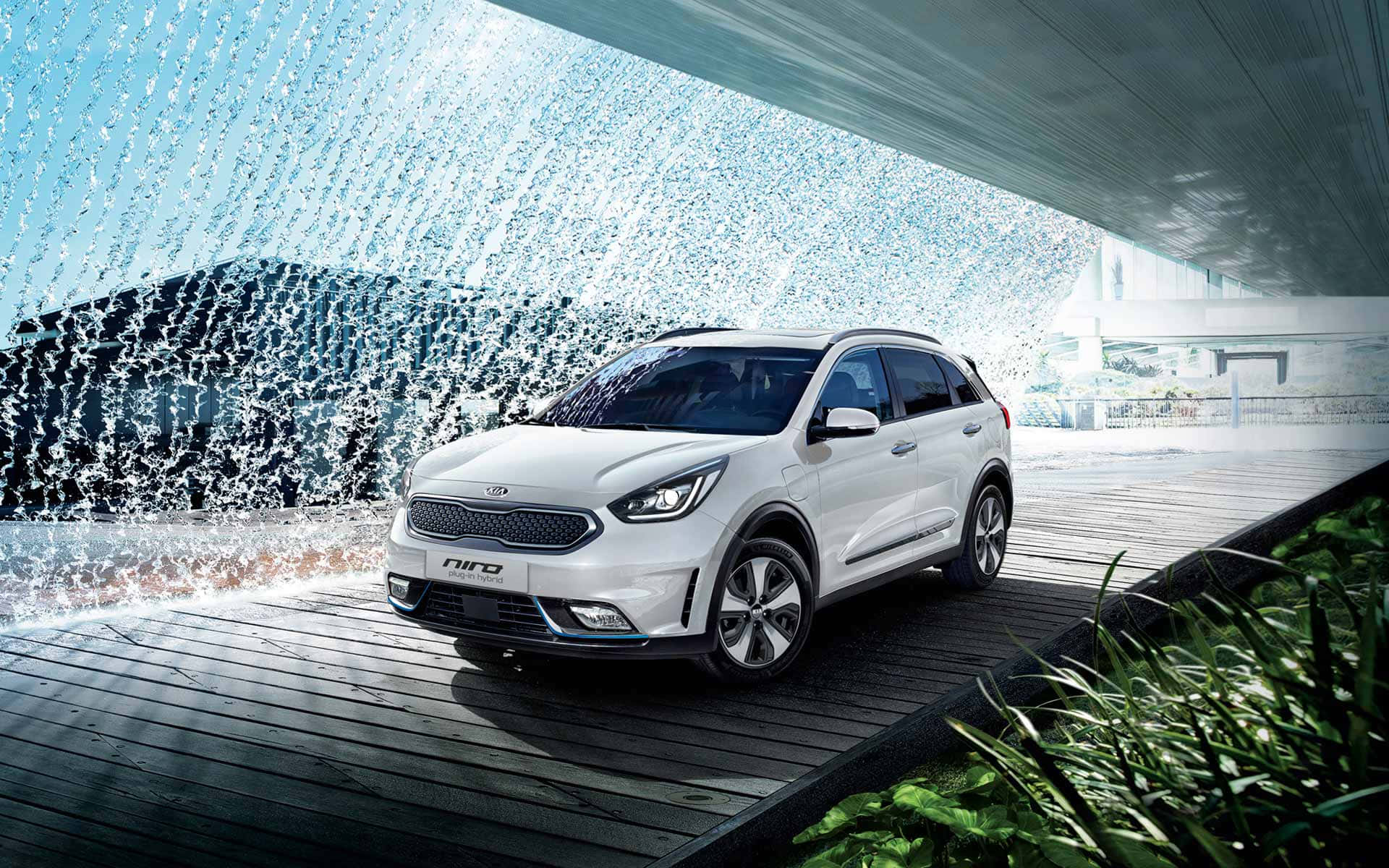 Kia Niro 桌布 壁紙 — 一輛時尚的白色Kia Niro SUV停放在現代建築簷篷下，附近有瀑布和綠植。