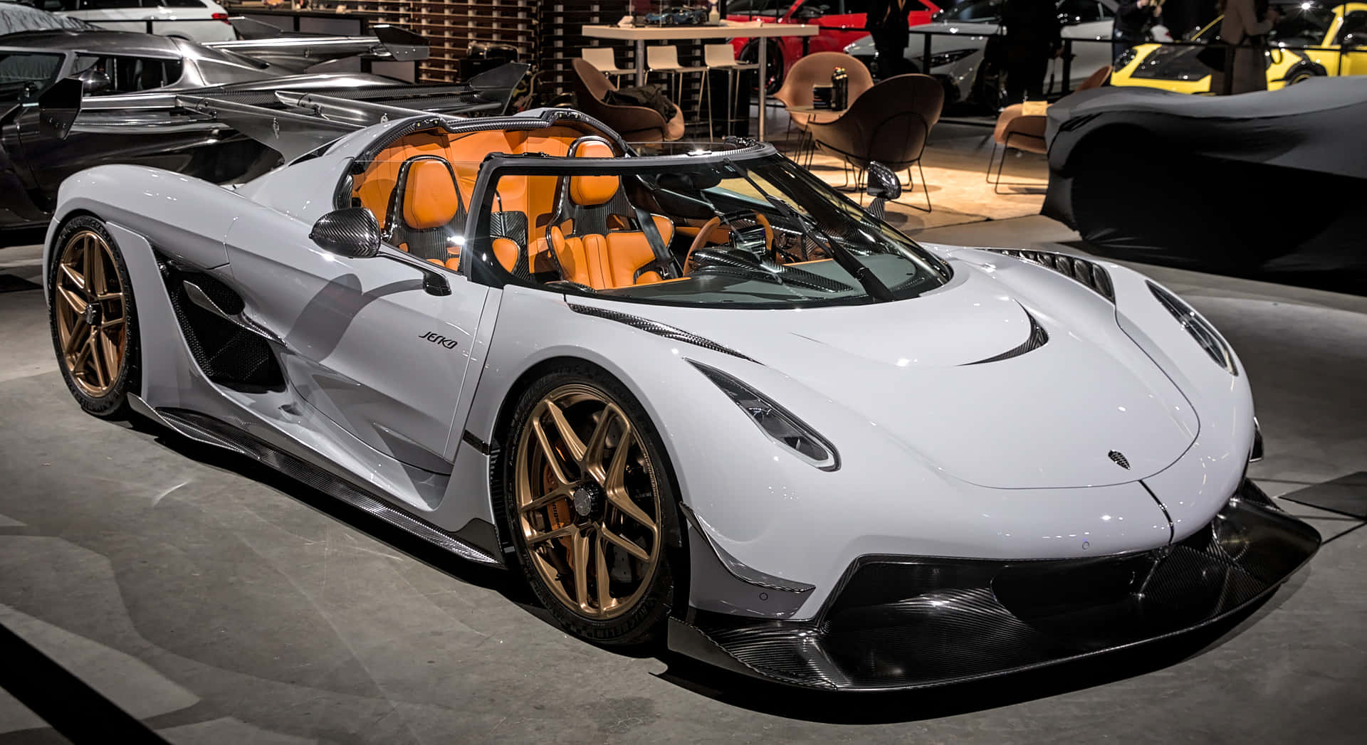  壁紙 — 一輛時尚的白色Koenigsegg Jesko，配備橙色內飾和金色輪圈，在車展的明亮燈光下閃閃發光。